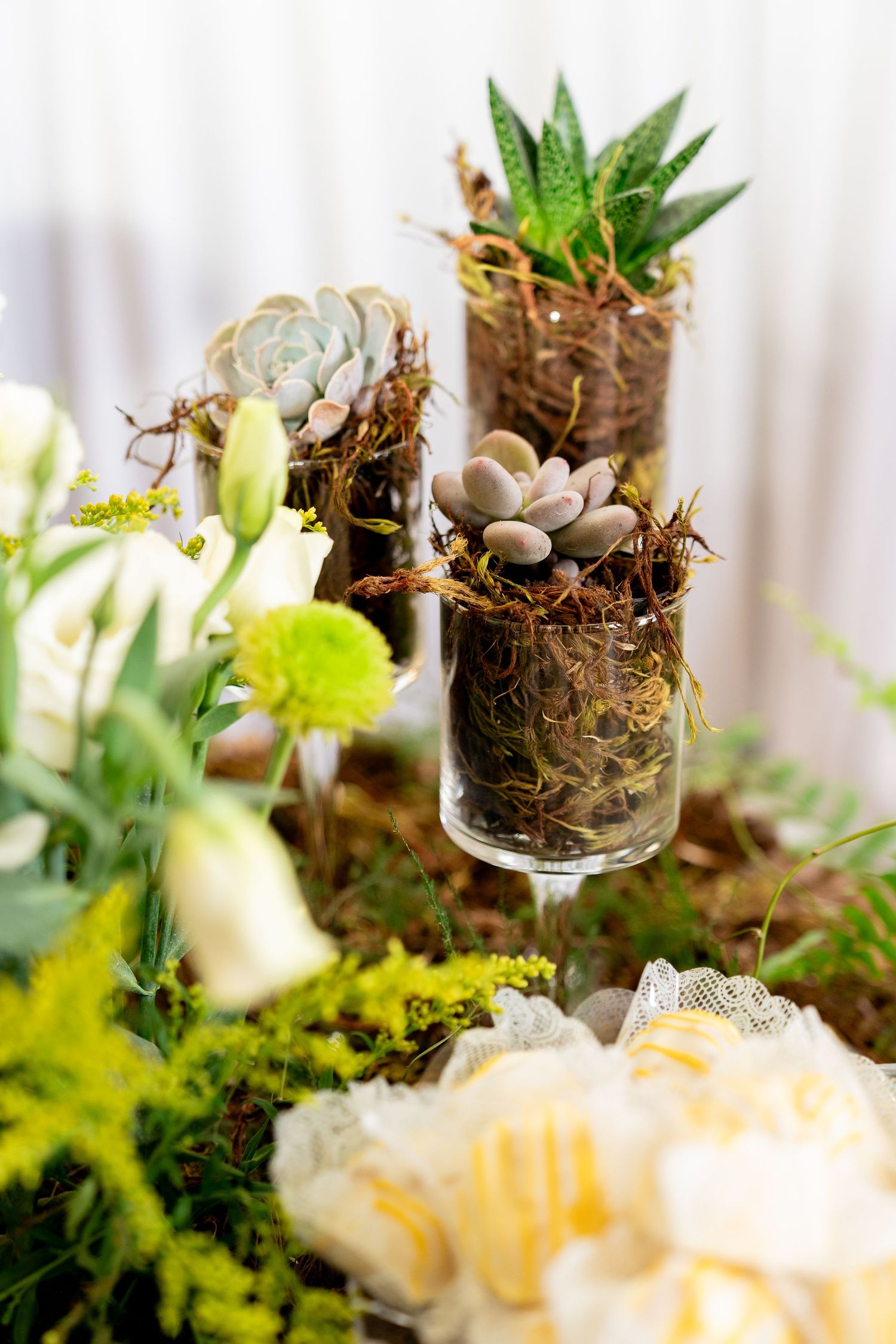 Glass vases with succulents and moss surrounded by white roses and yellow flowers for elegant table decor.