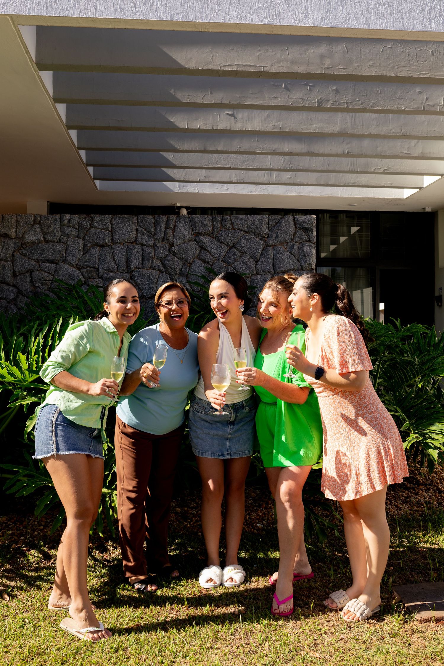 Five women smiling and holding champagne glasses outdoors in front of a modern building with lush greenery.