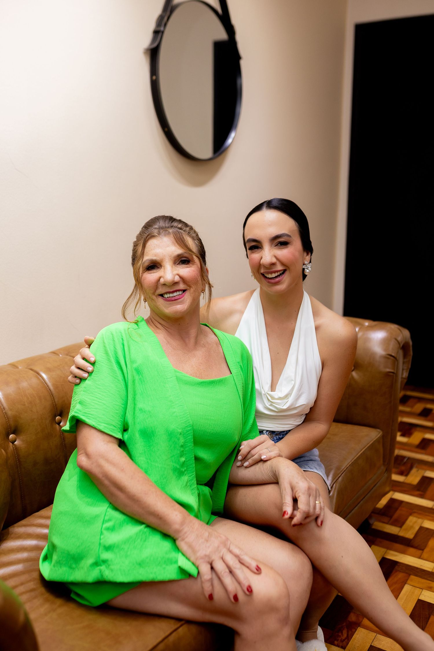 Two women smiling and posing on a brown leather sofa, one in green dress, other in white top and denim shorts.
