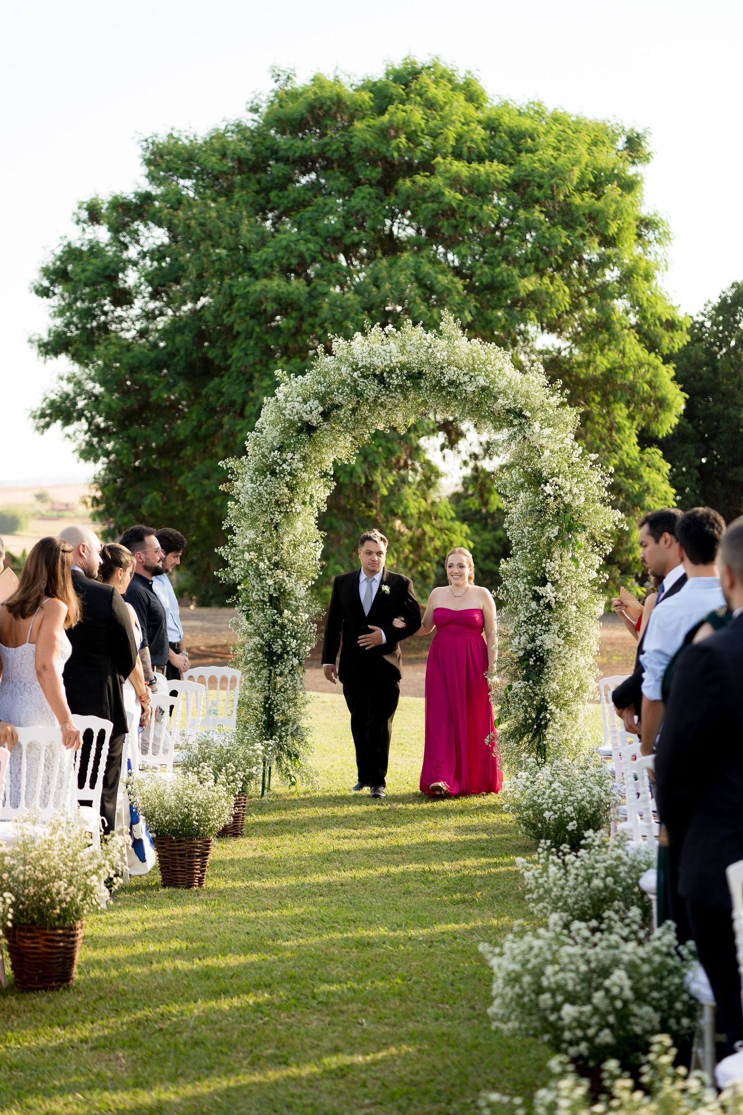 Outdoor wedding ceremony with floral arch, groom in tuxedo, bridesmaid in pink gown, guests seated on white chairs.