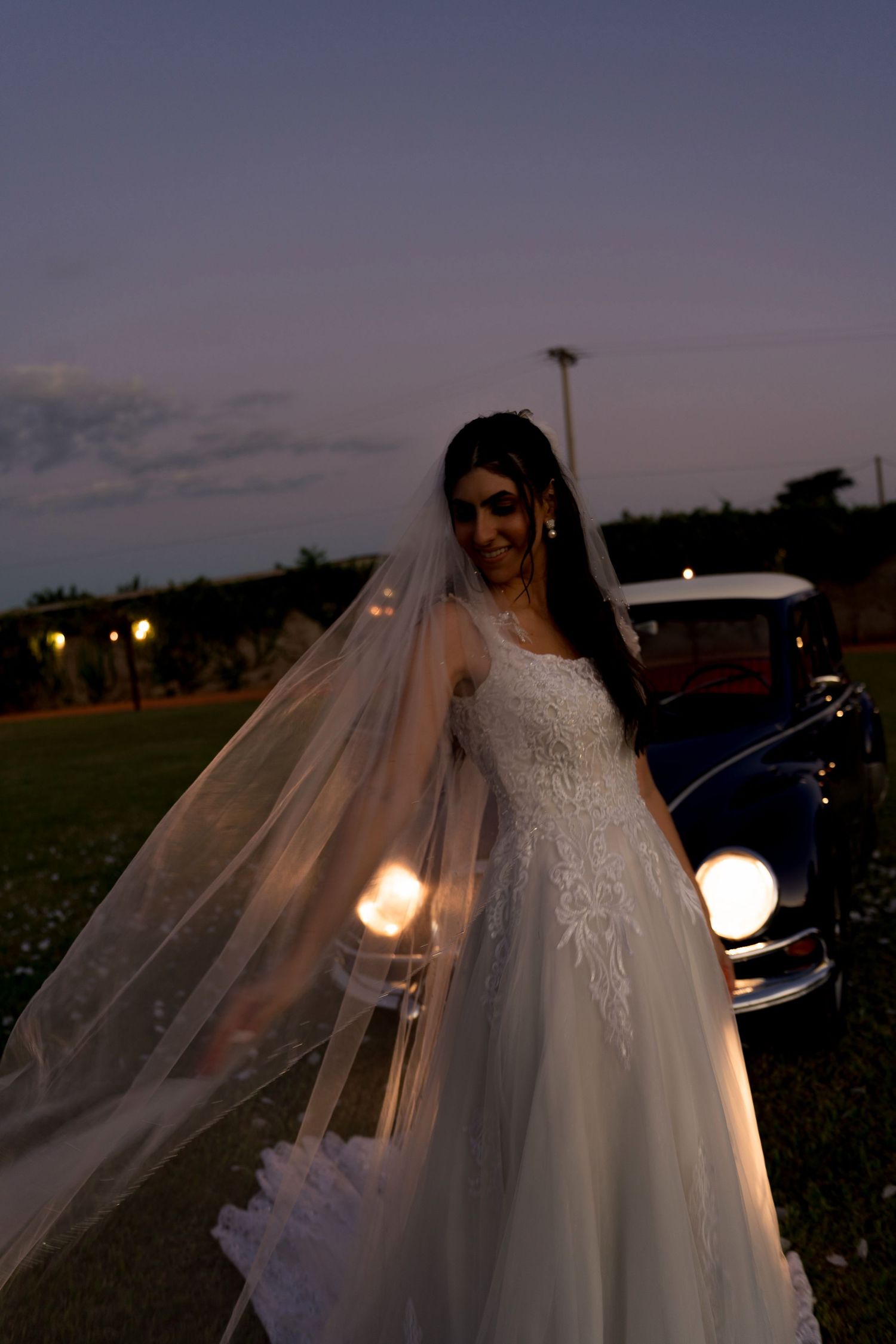 Bride in lace wedding gown and veil poses outdoors at dusk near a vintage car with glowing headlights.