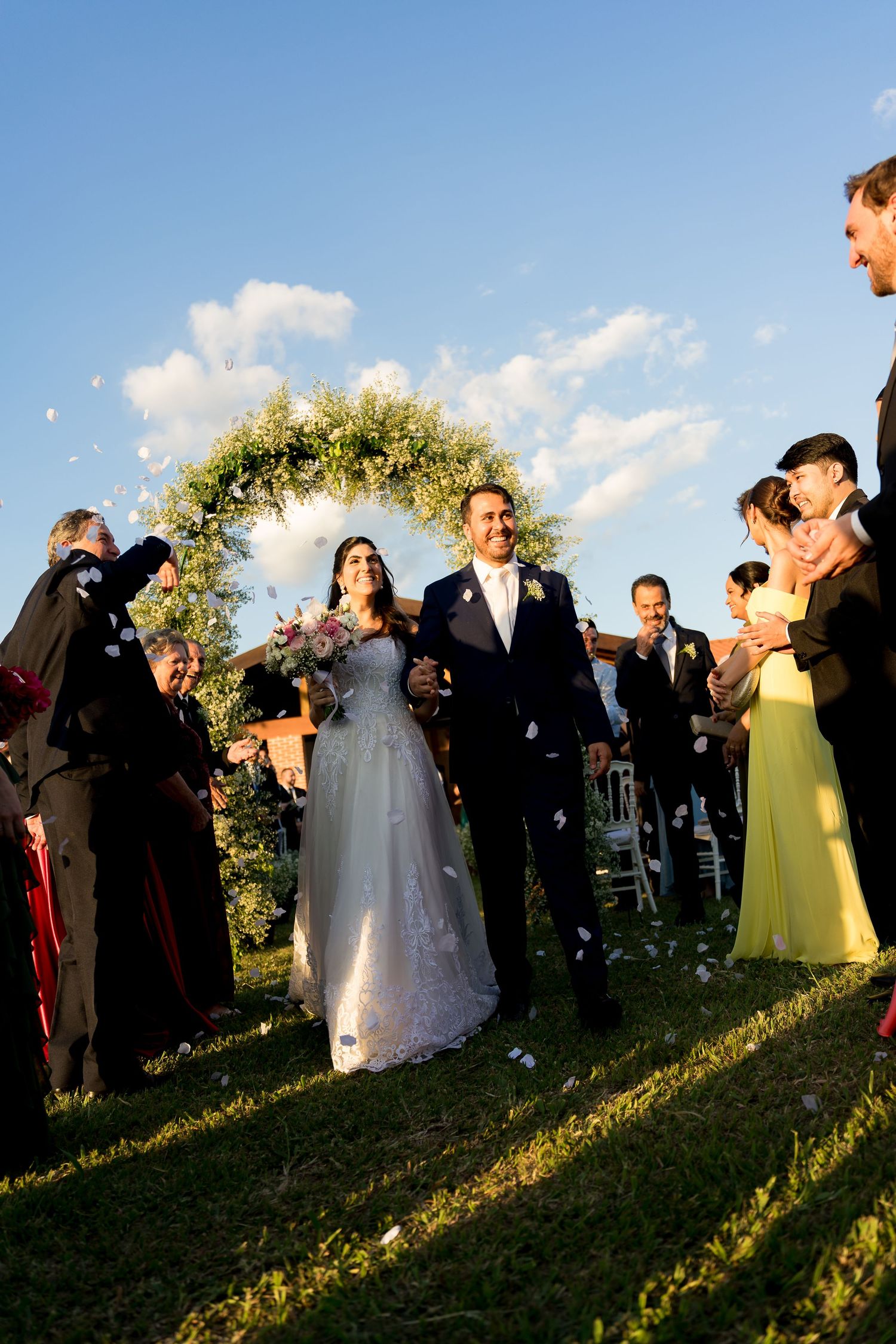 Bride and groom walk down the aisle as guests toss flower petals at an outdoor wedding ceremony under a blue sky.