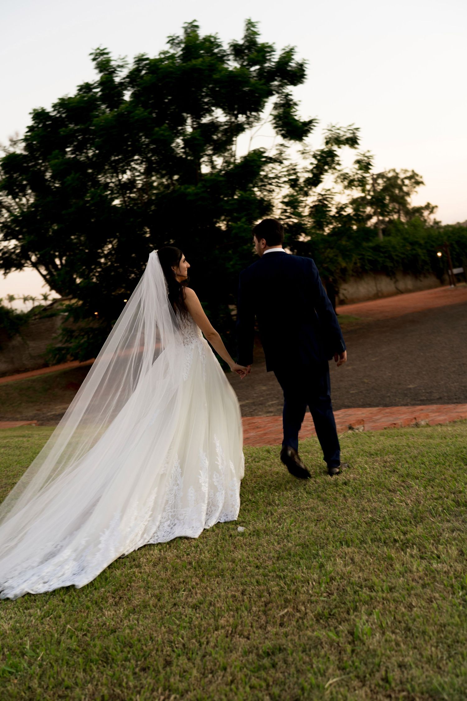 Bride in white gown with long veil walks hand in hand with groom in navy suit at dusk near lush green trees.