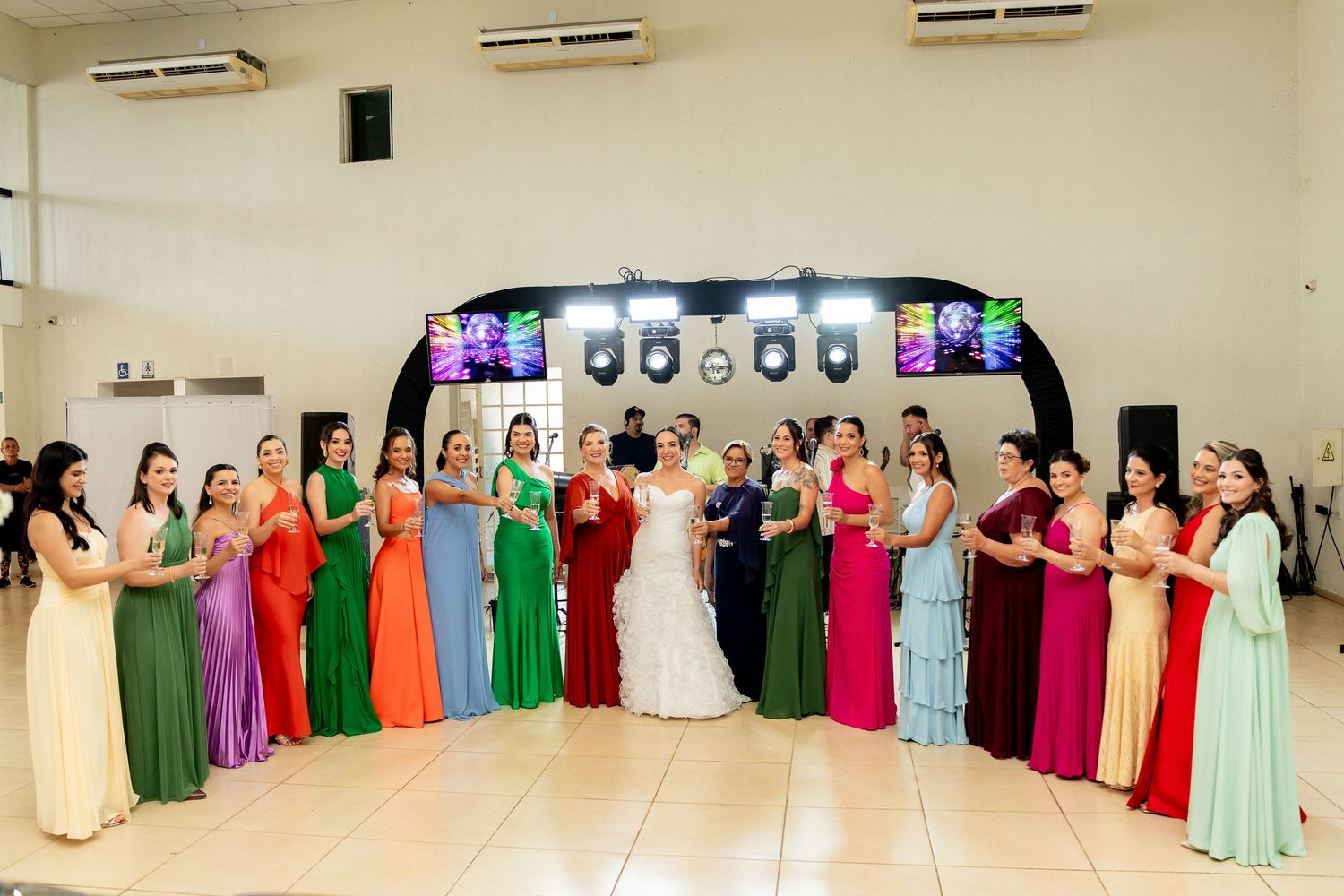 Bride surrounded by bridesmaids in colorful rainbow dresses posing together on a dance floor at a wedding reception.