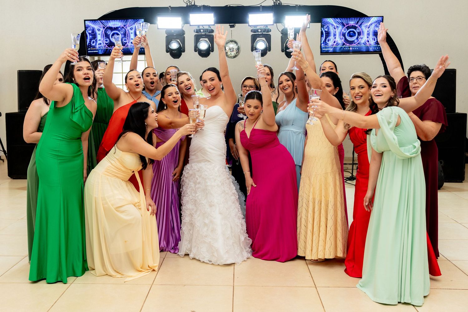 Bride in white gown poses with colorful bridesmaids in rainbow dresses, celebrating joyfully at a wedding reception.