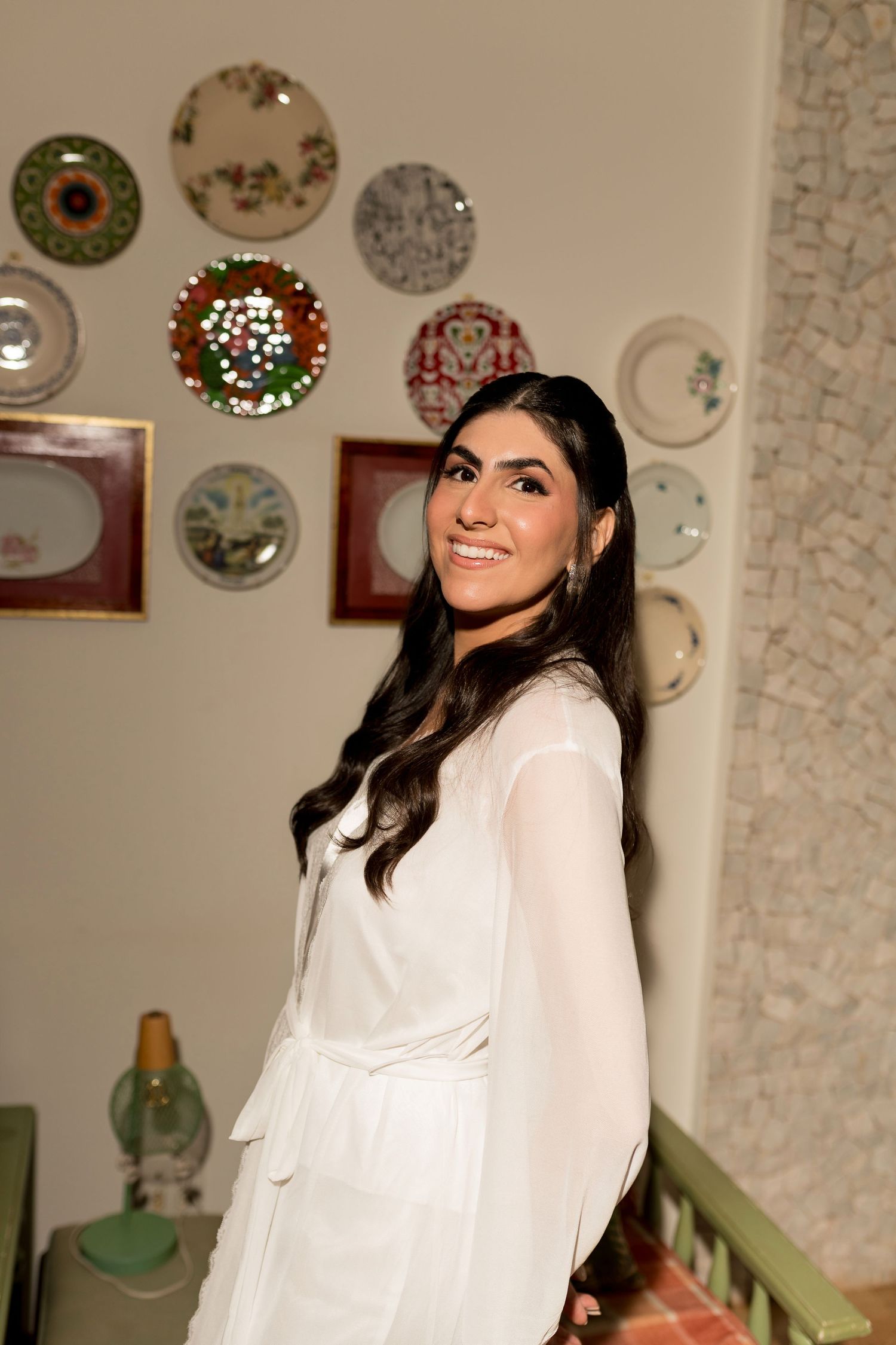 Smiling woman in white outfit poses indoors with decorative plates displayed on wall behind her.