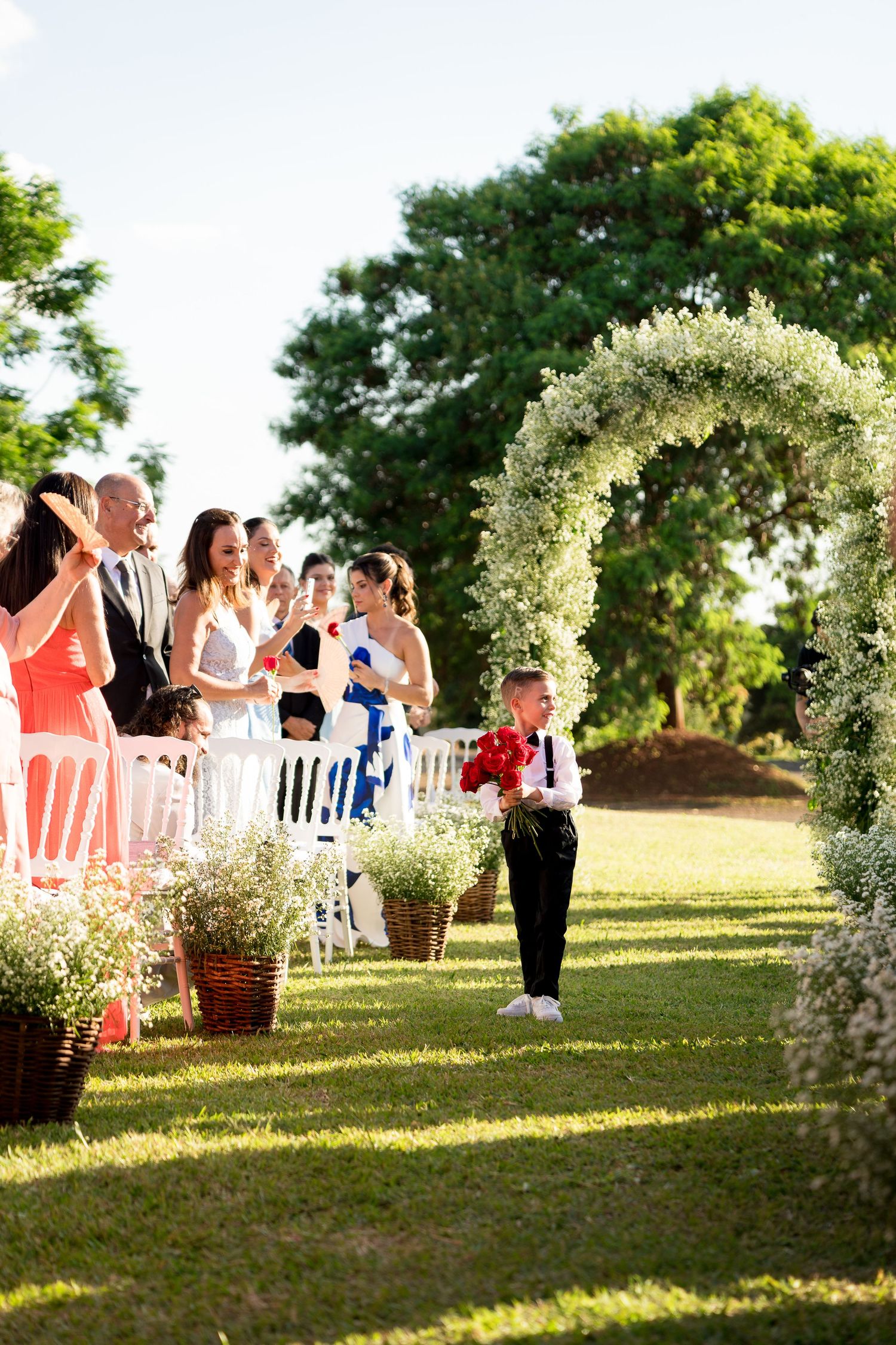 Young ring bearer walks down outdoor wedding aisle under floral arch, guests seated on white chairs, lush green trees backdrop.