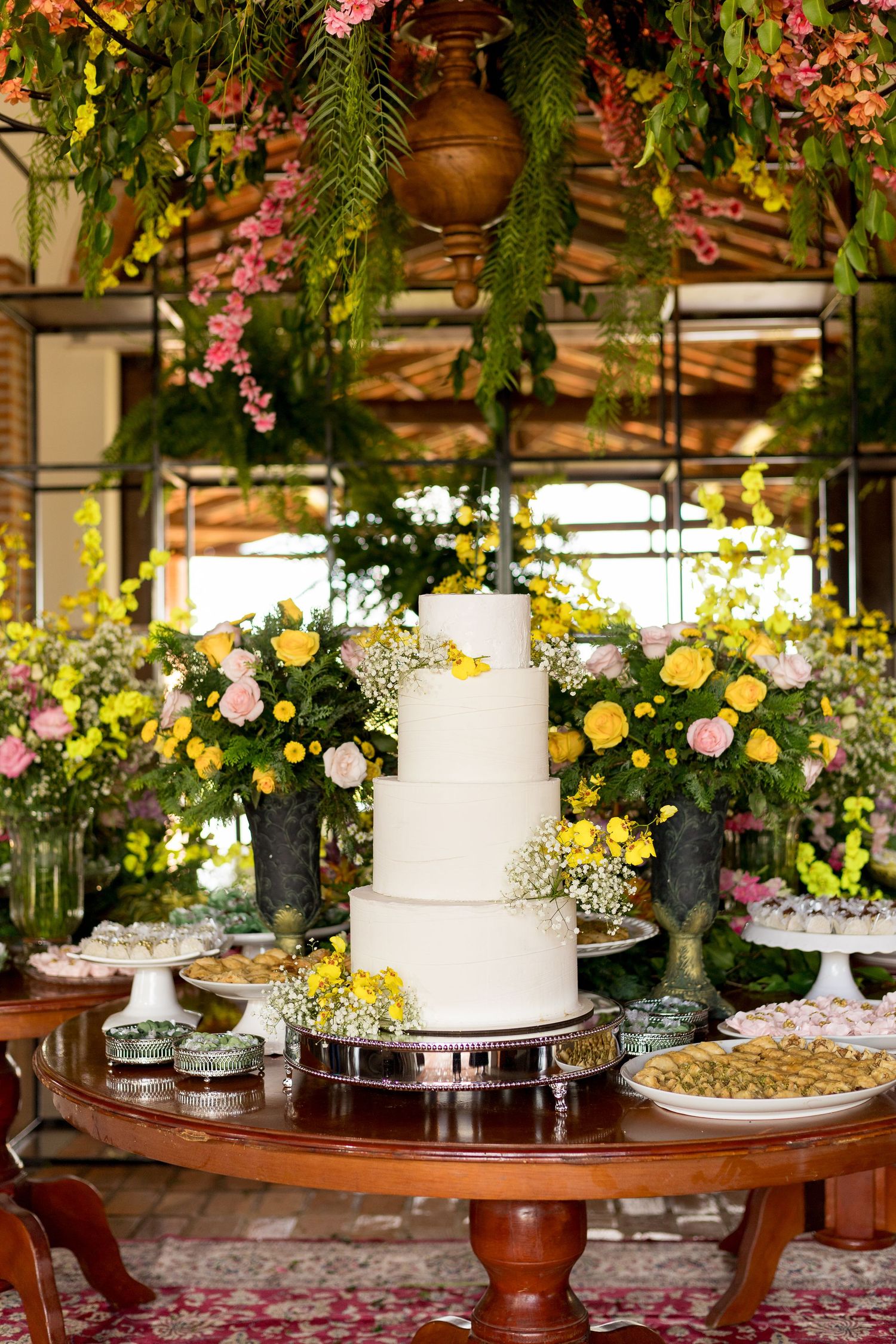 Elegant 4-tier white wedding cake on wooden table surrounded by yellow and pink floral arrangements in lush garden setting.