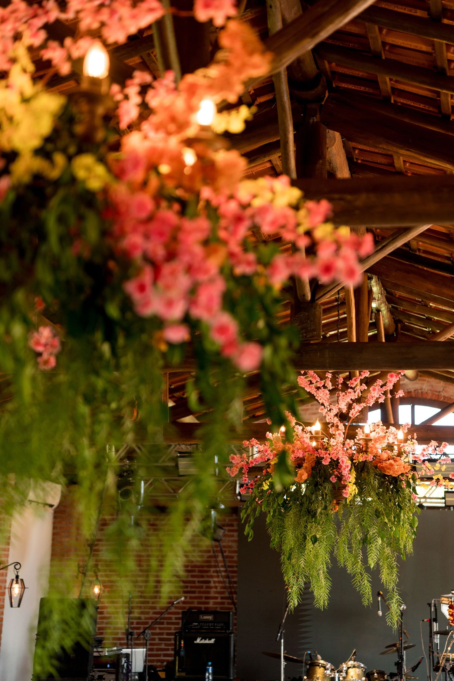 Lush pink and green floral arrangements hang from rustic wooden ceiling beams in a warmly lit barn venue.