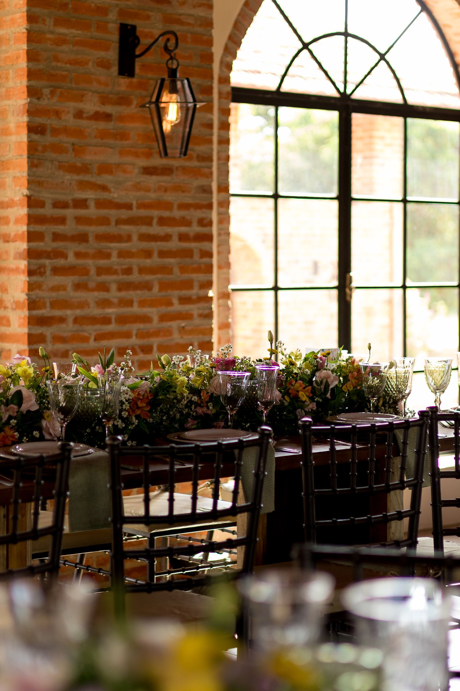 Elegant rustic wedding reception venue with brick walls, arched windows, floral centerpieces, and dark chiavari chairs set for dining.