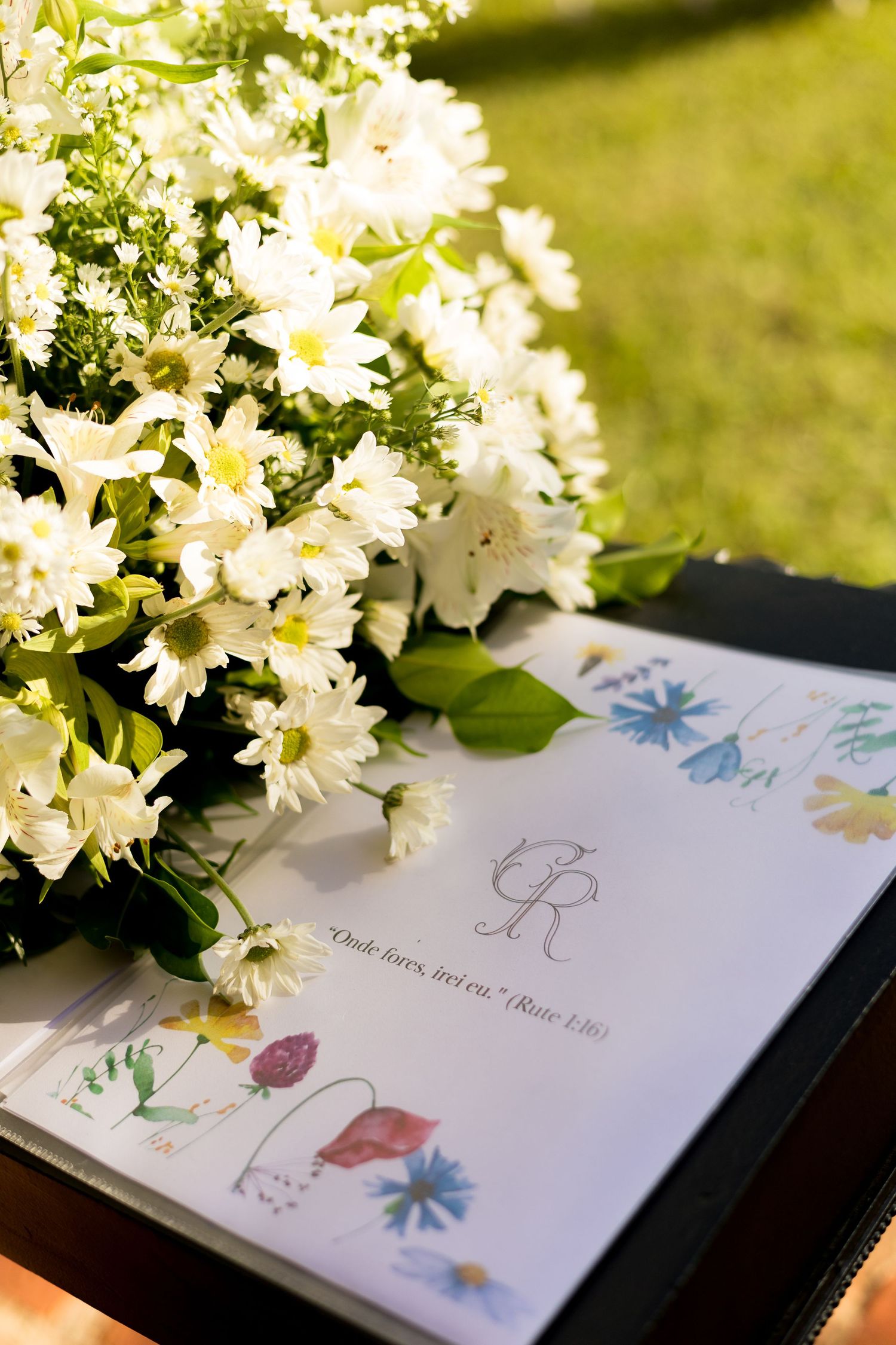 White floral bouquet with daisies beside an illustrated wedding program featuring monogram and botanical drawings on a dark surface.