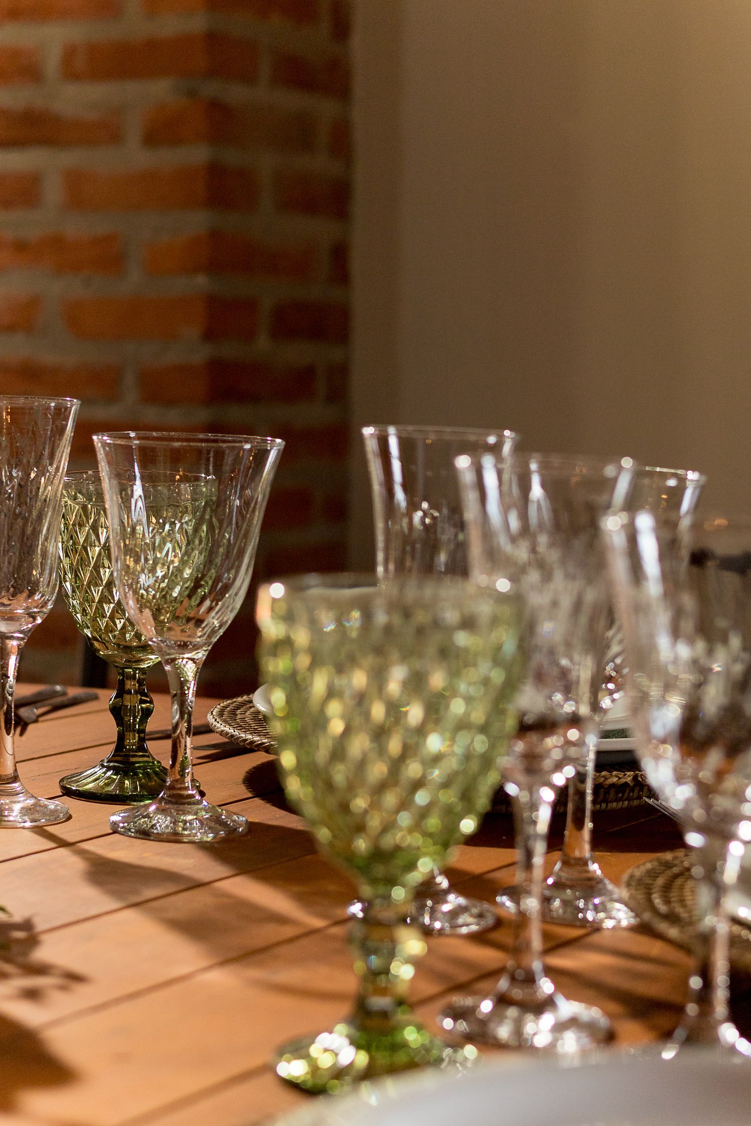 Elegant crystal wine glasses in clear and green hues arranged on a wooden table near a brick wall.