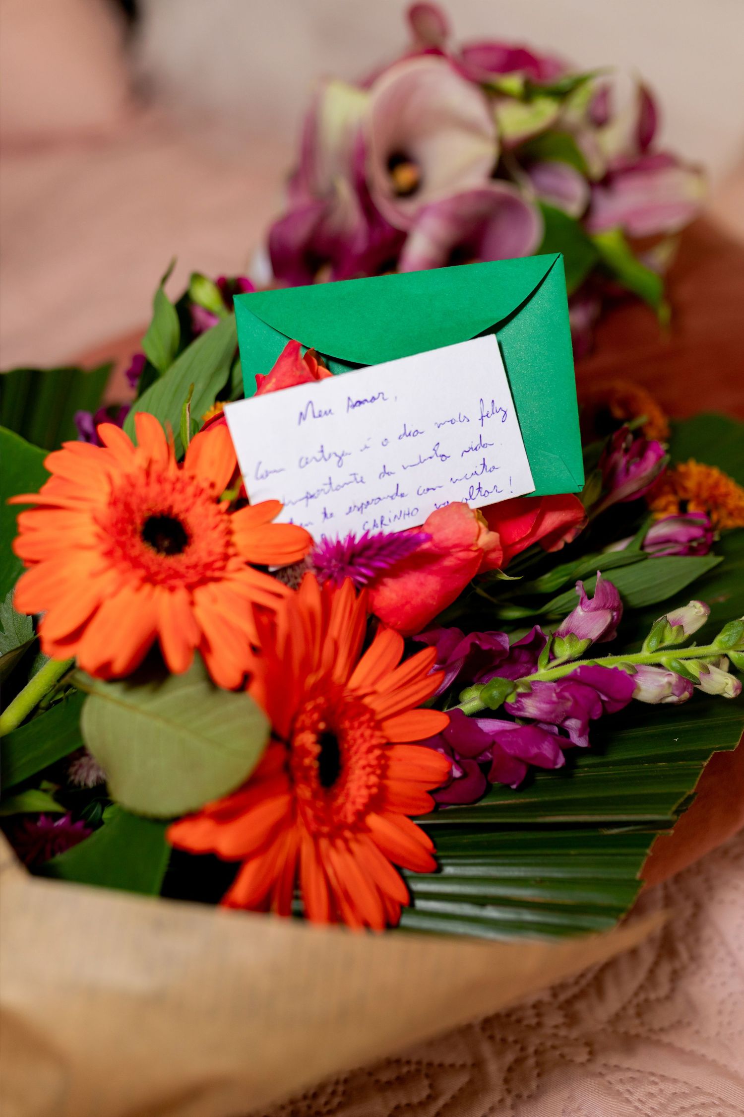 Colorful bouquet of orange gerbera daisies and purple flowers with a handwritten note in a green envelope.