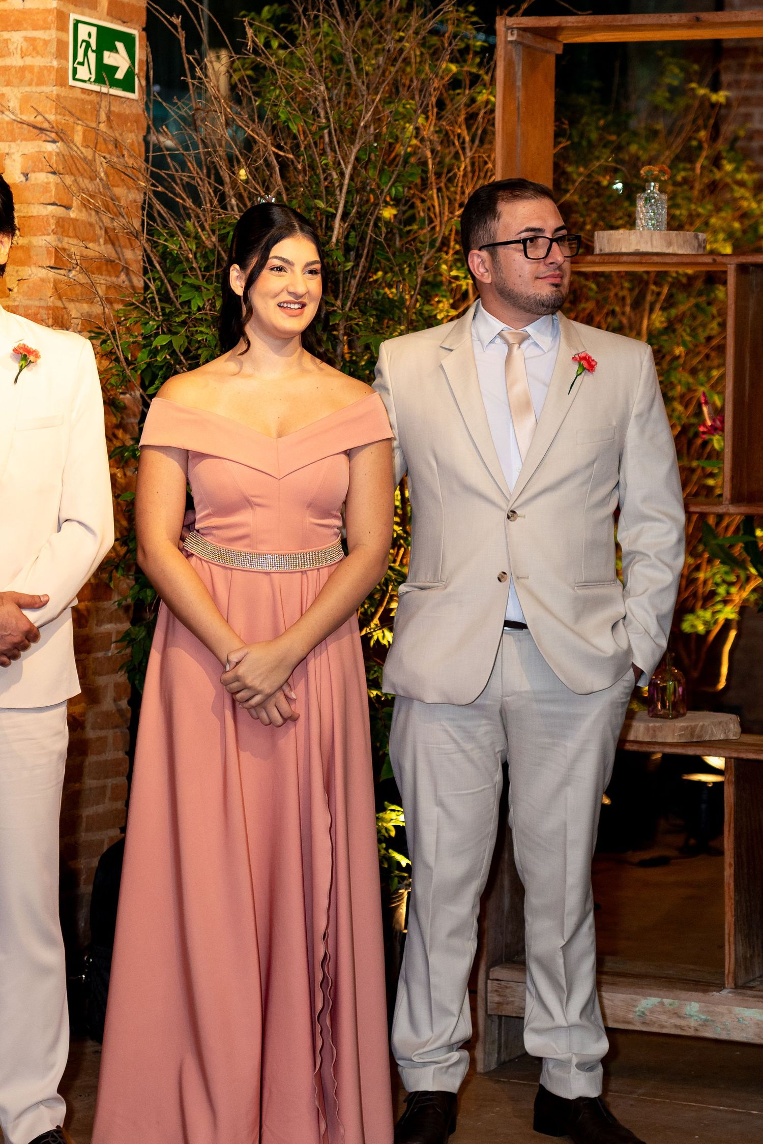 Couple in formal attire at a wedding, woman in pink off-shoulder gown, man in beige suit with boutonniere.