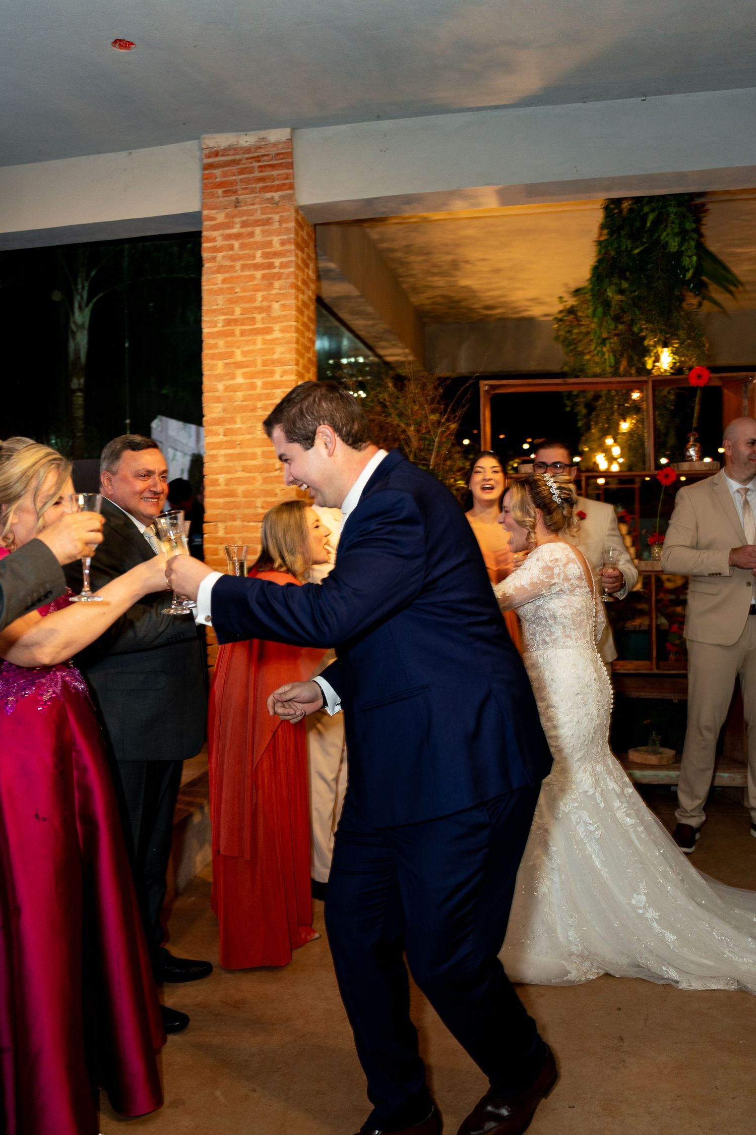 Bride and groom dancing joyfully at wedding reception surrounded by guests in elegant venue with brick wall backdrop.