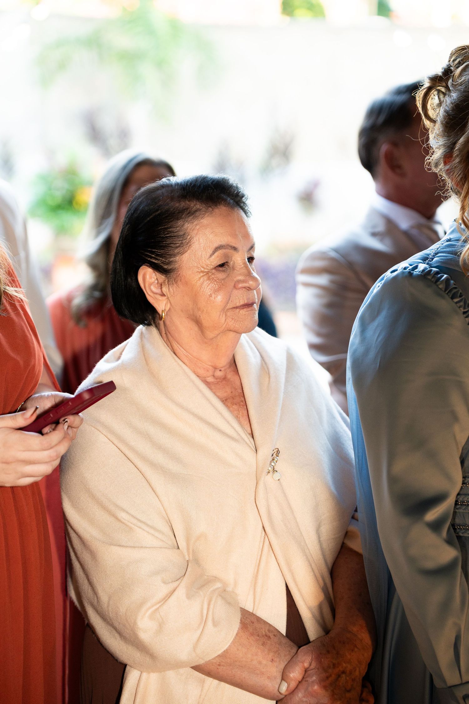 Elderly woman in white shawl with brooch attends a formal event, smiling graciously among other guests.