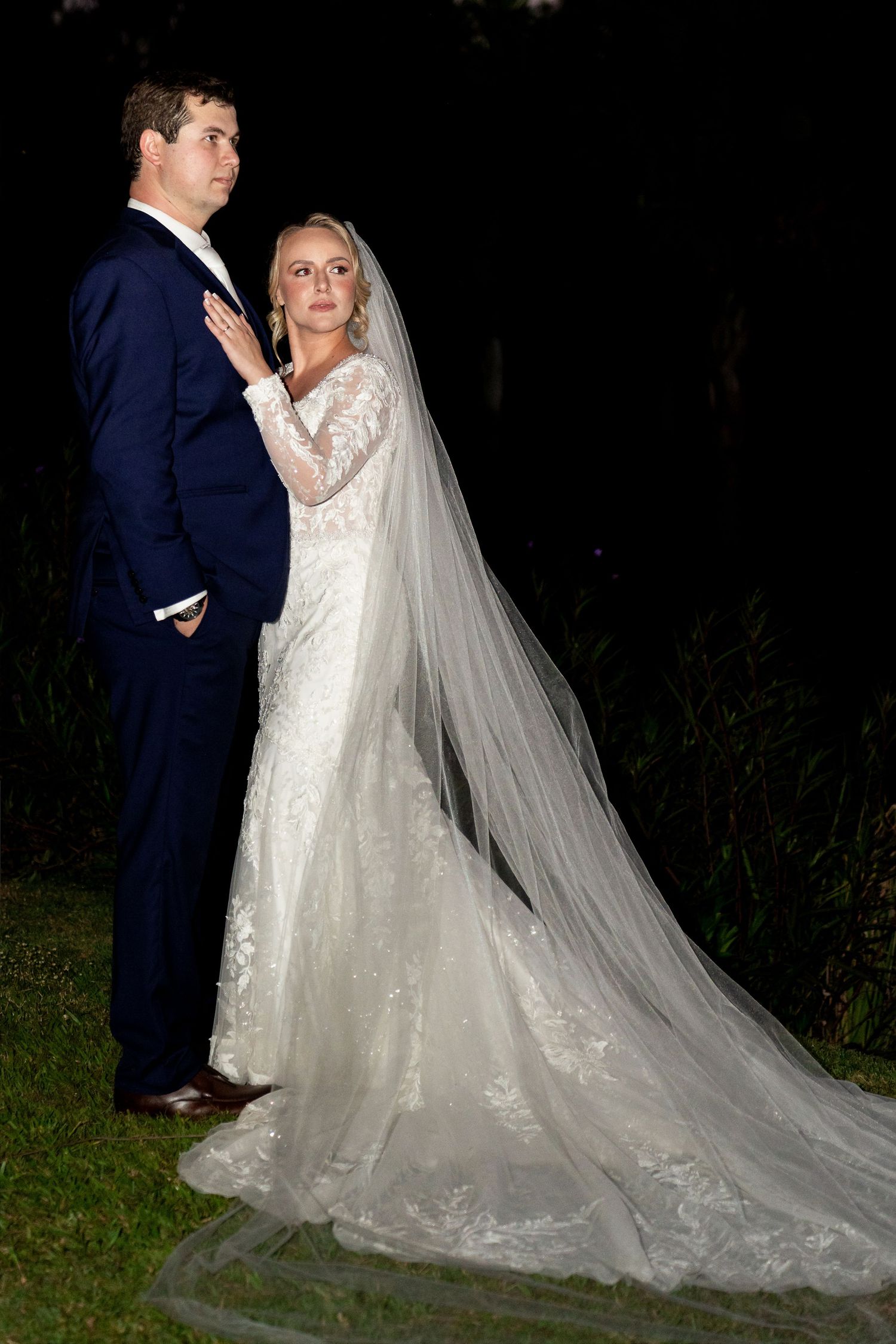 Bride in lace gown with cathedral veil stands beside groom in navy suit during outdoor evening wedding portrait.