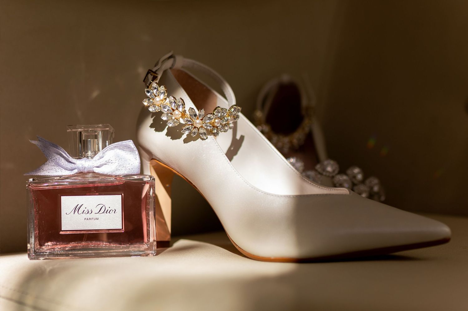 White bridal heels with jeweled ankle strap beside Miss Dior perfume bottle on golden surface.