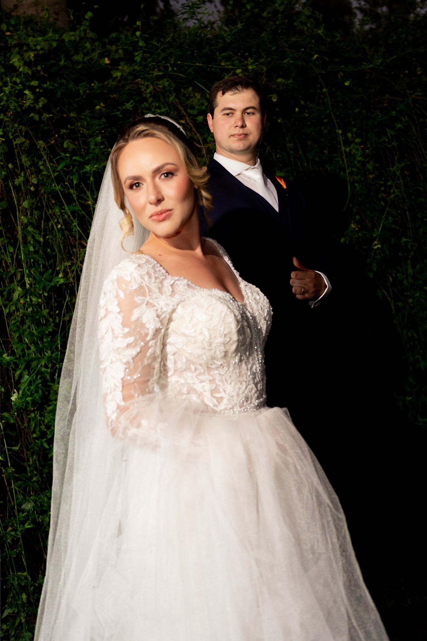 Bride in lace gown and veil with groom in black tuxedo posing together outdoors at night.