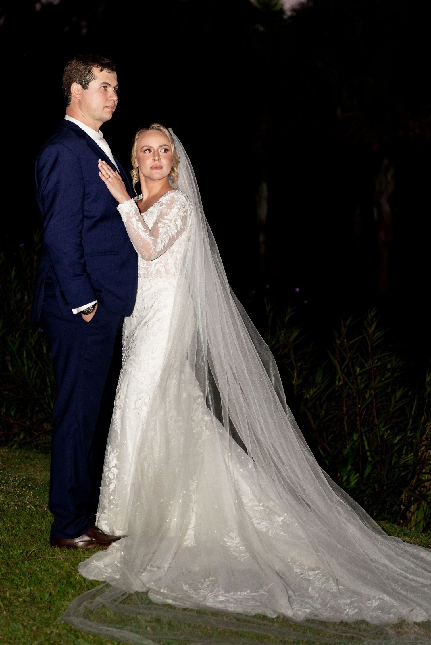 A bride in a lace gown with cathedral veil and groom in navy suit pose elegantly outdoors at night.