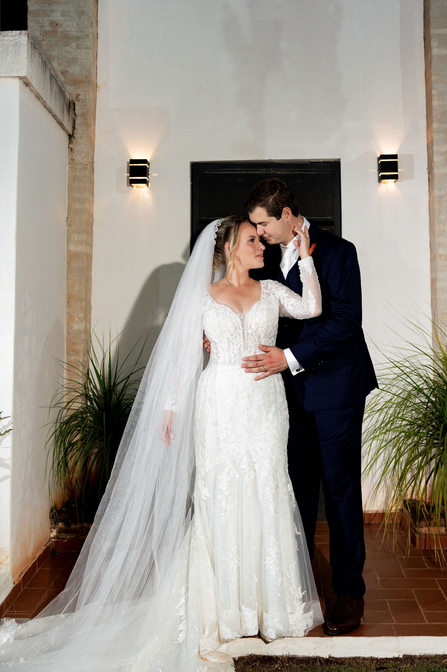 Bride in lace gown with veil and groom in navy suit share romantic moment at elegant evening wedding venue.