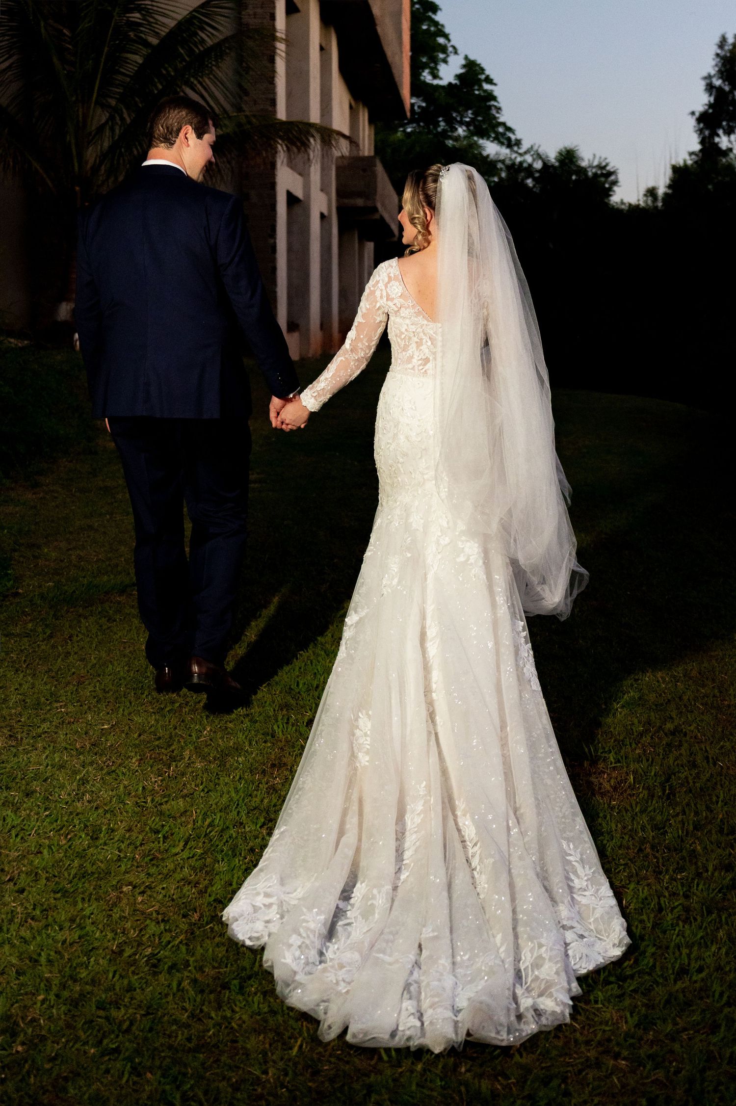 Bride in lace long-sleeve mermaid gown with veil holds hands with groom in navy suit walking outdoors at golden hour.