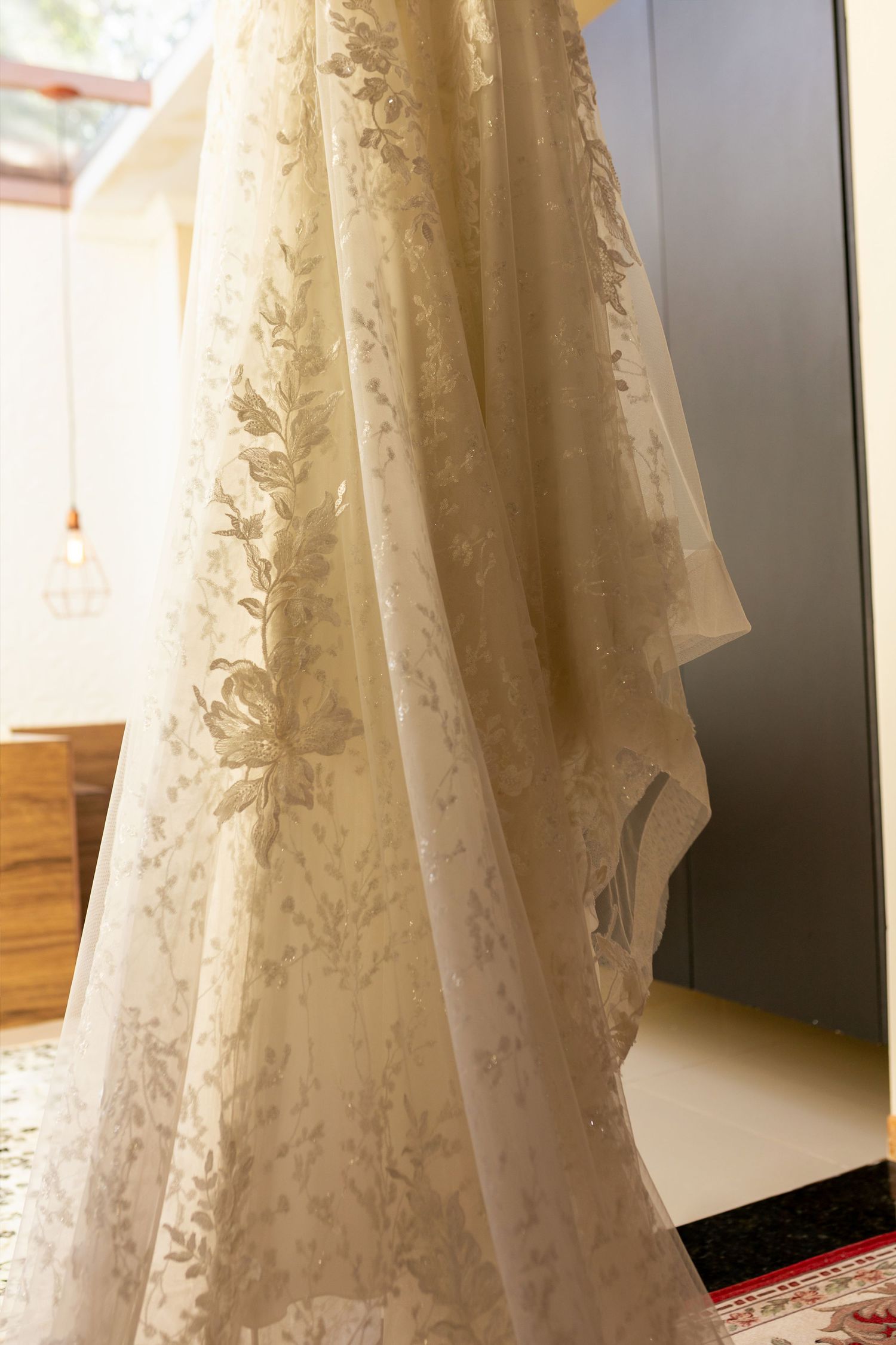 Elegant ivory lace wedding dress with floral embroidery hanging near a mirror in a softly lit bridal suite.
