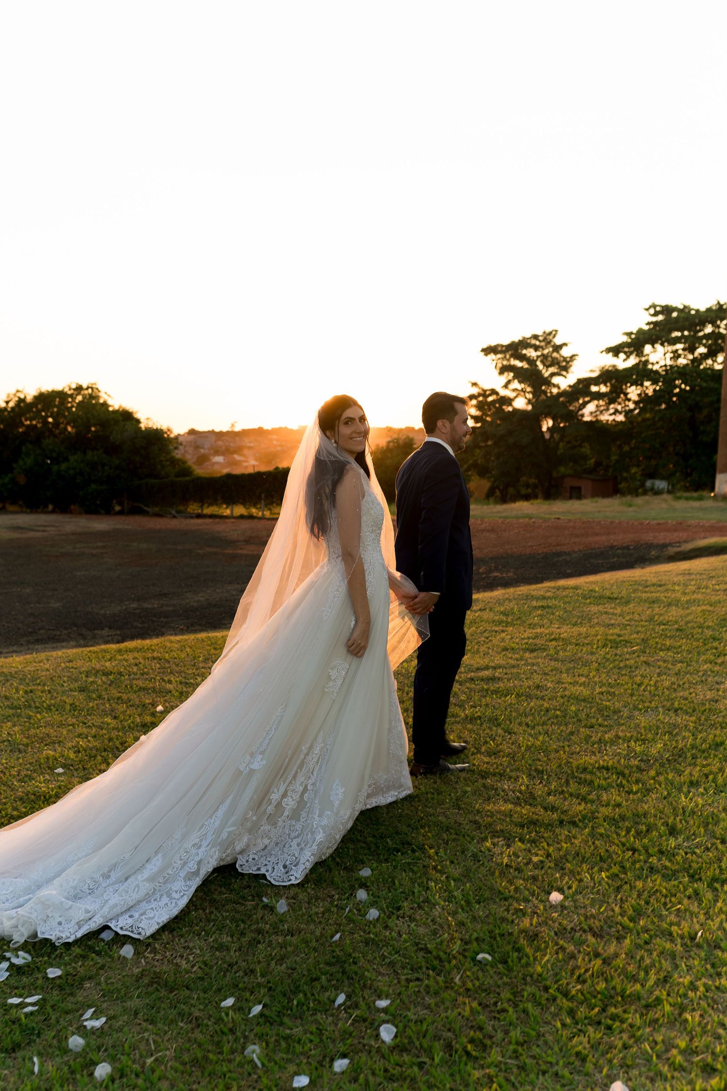 Bride in lace gown and veil holds groom's hand at sunset on green lawn with trees in background.