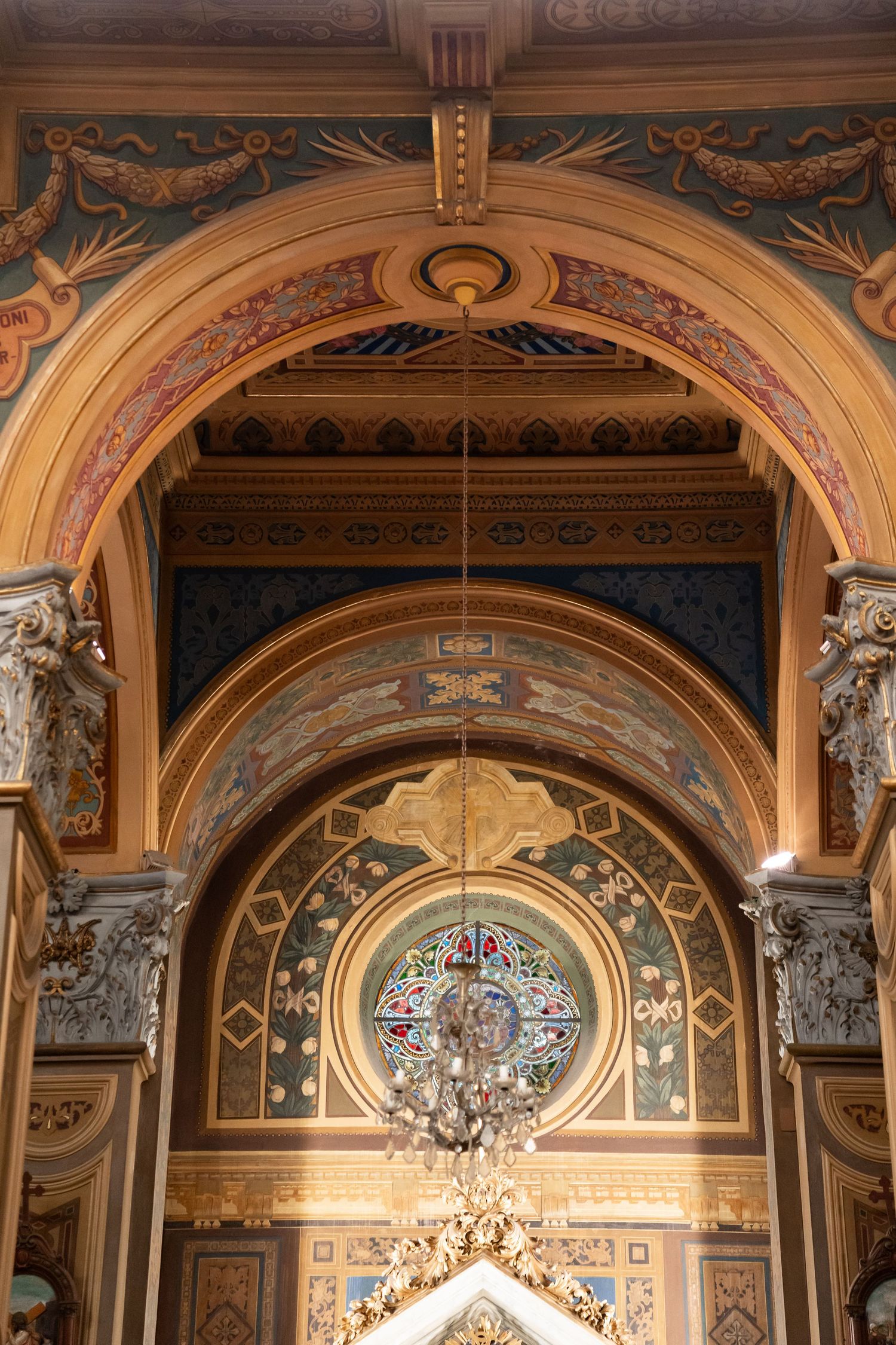 Ornate cathedral interior with gilded arches, colorful frescoes, stained glass rose window, and elegant chandelier.