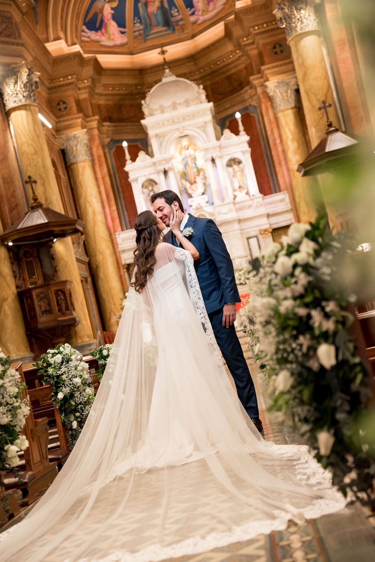 Bride and groom share a romantic kiss at the altar of an ornate church, with floral arrangements and a long veil.