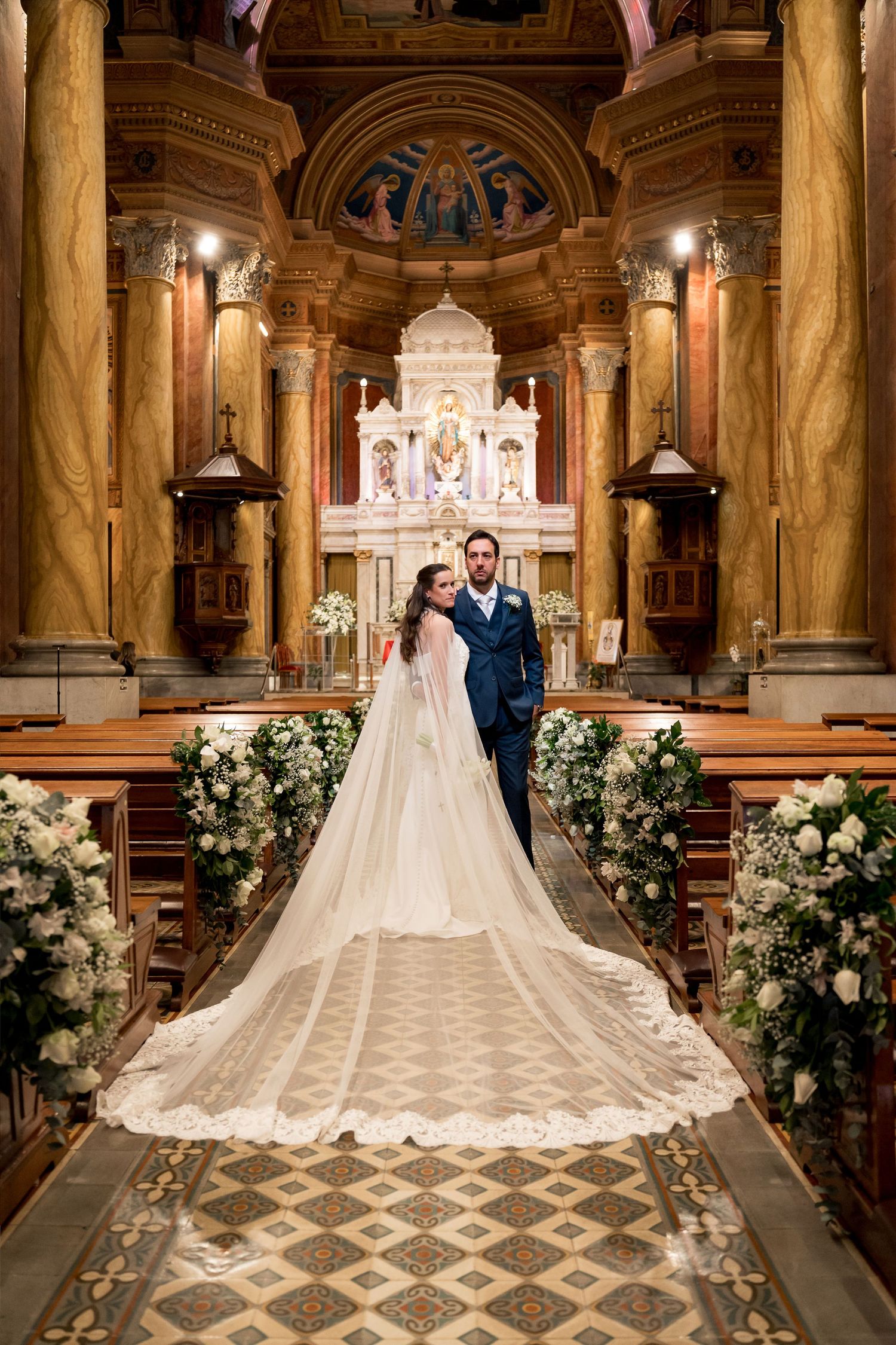 Bride and groom stand at church altar, bride's lace cathedral veil trailing down ornate aisle lined with white floral arrangements.