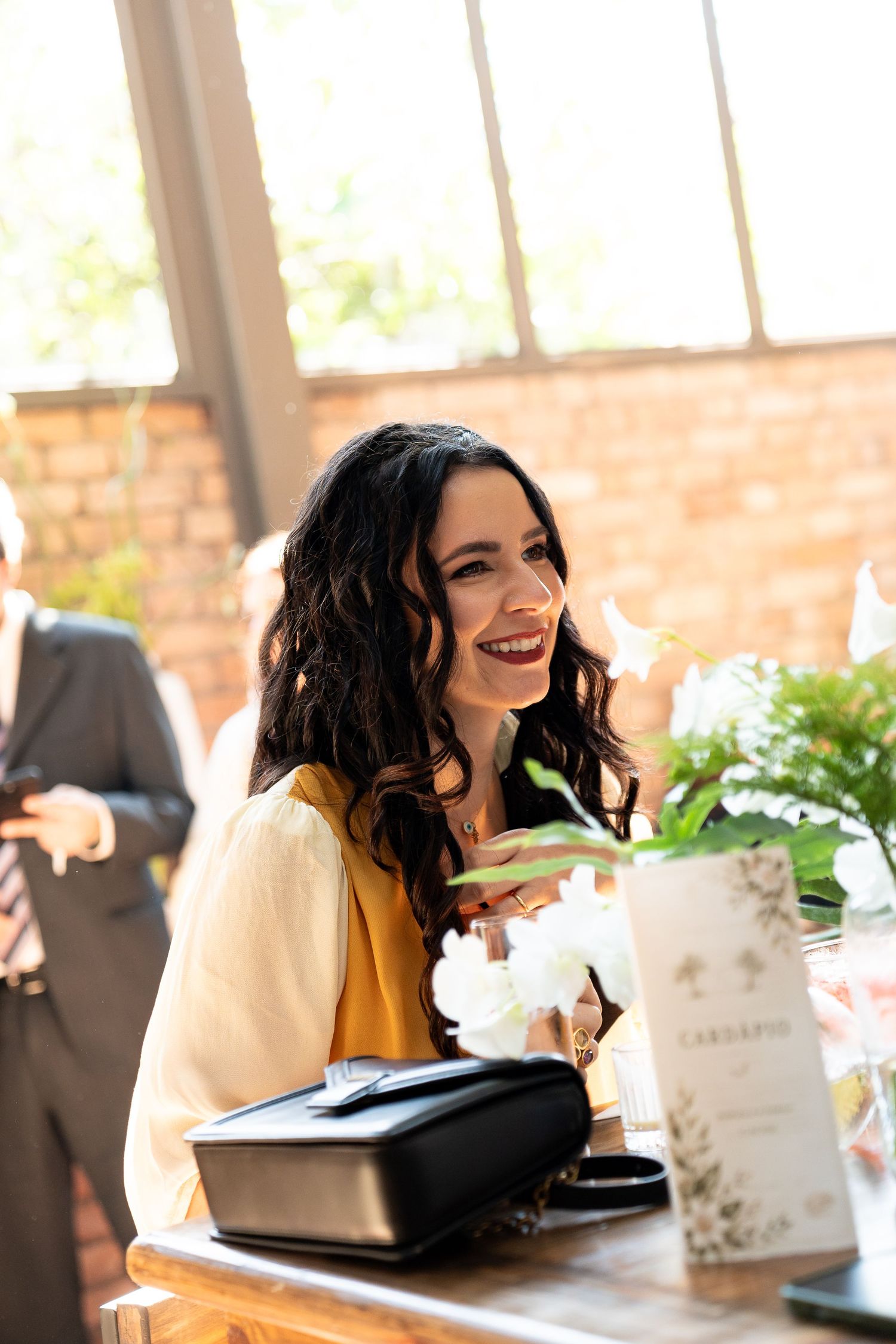 Smiling brunette woman in yellow dress seated at elegant wedding reception table with white flowers.