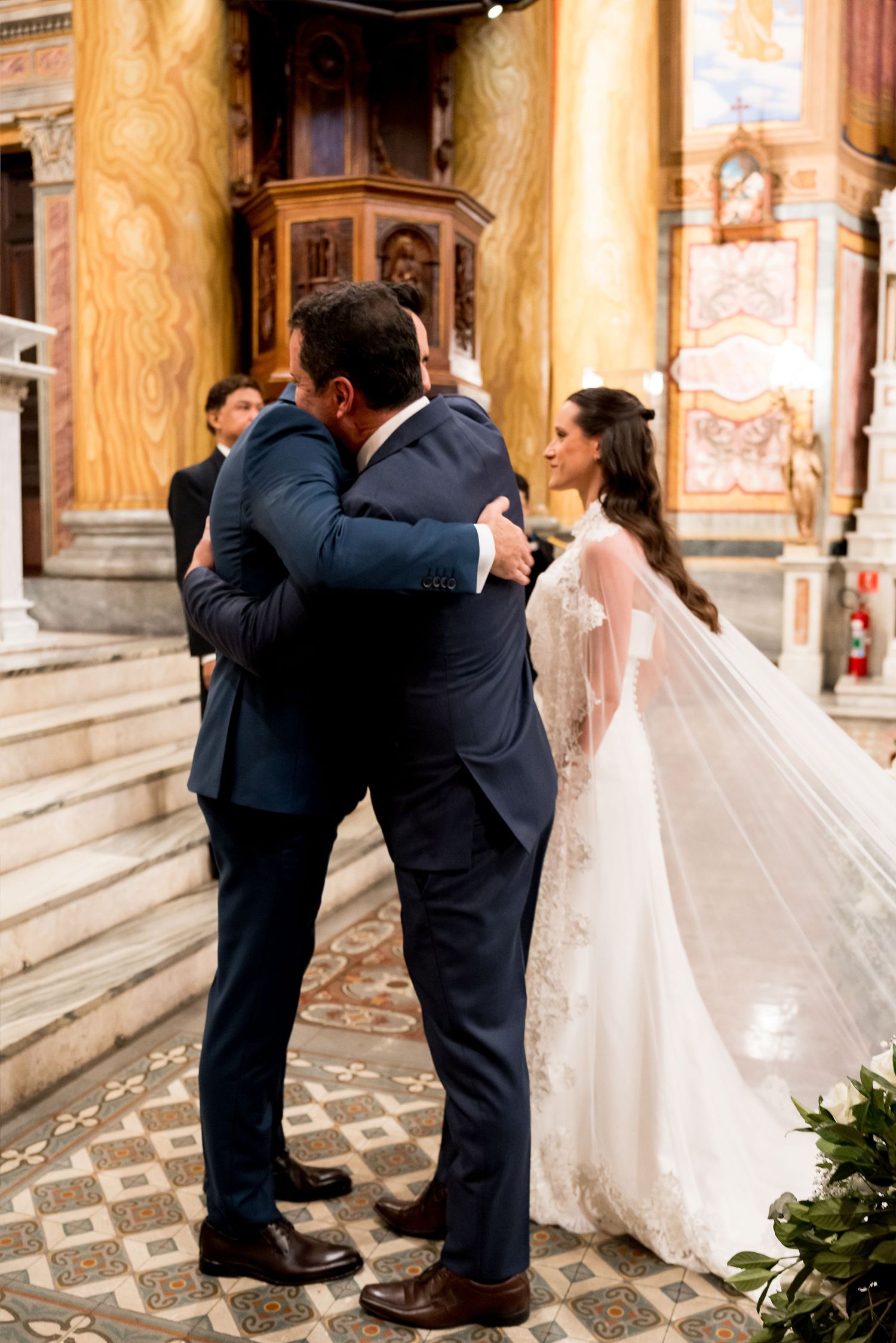 Groom embracing a guest in a church while bride in lace wedding gown and veil stands nearby smiling.