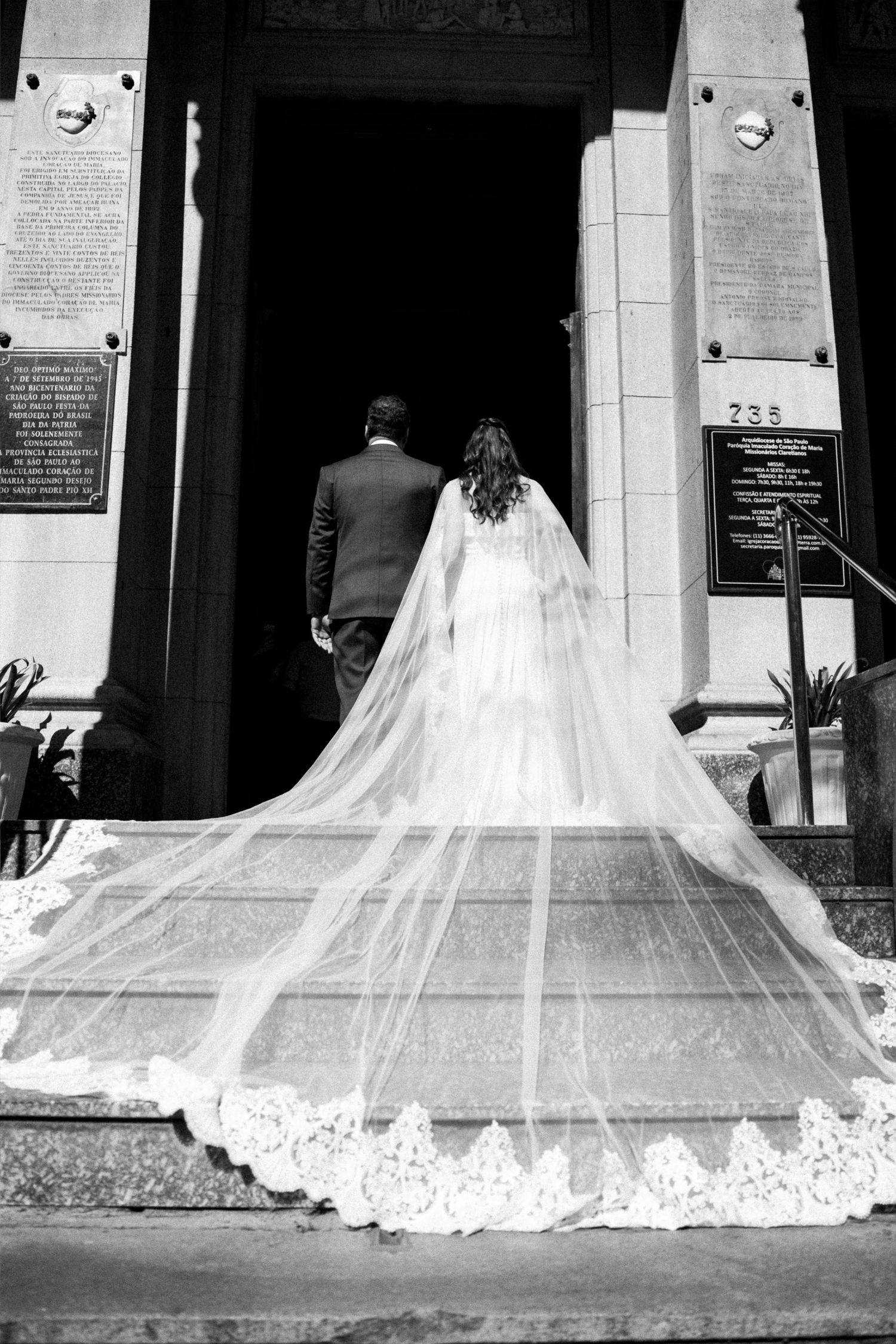 Black and white photo of bride and groom entering church, bride wearing long lace-trimmed cathedral veil.