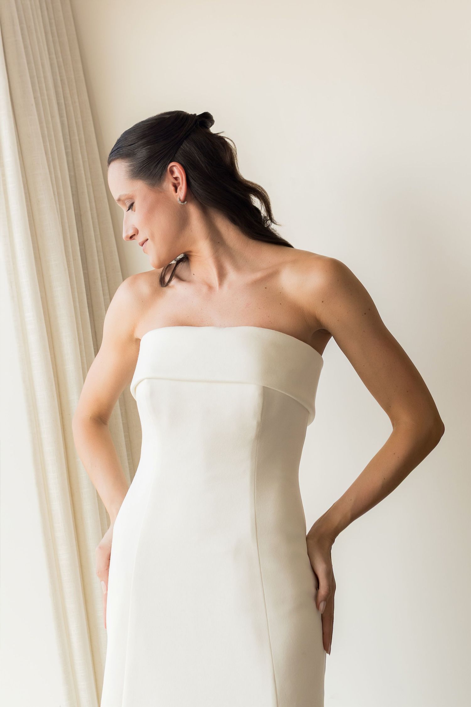 Woman in elegant strapless white gown with hands on hips, dark hair in ponytail, posing near curtain.