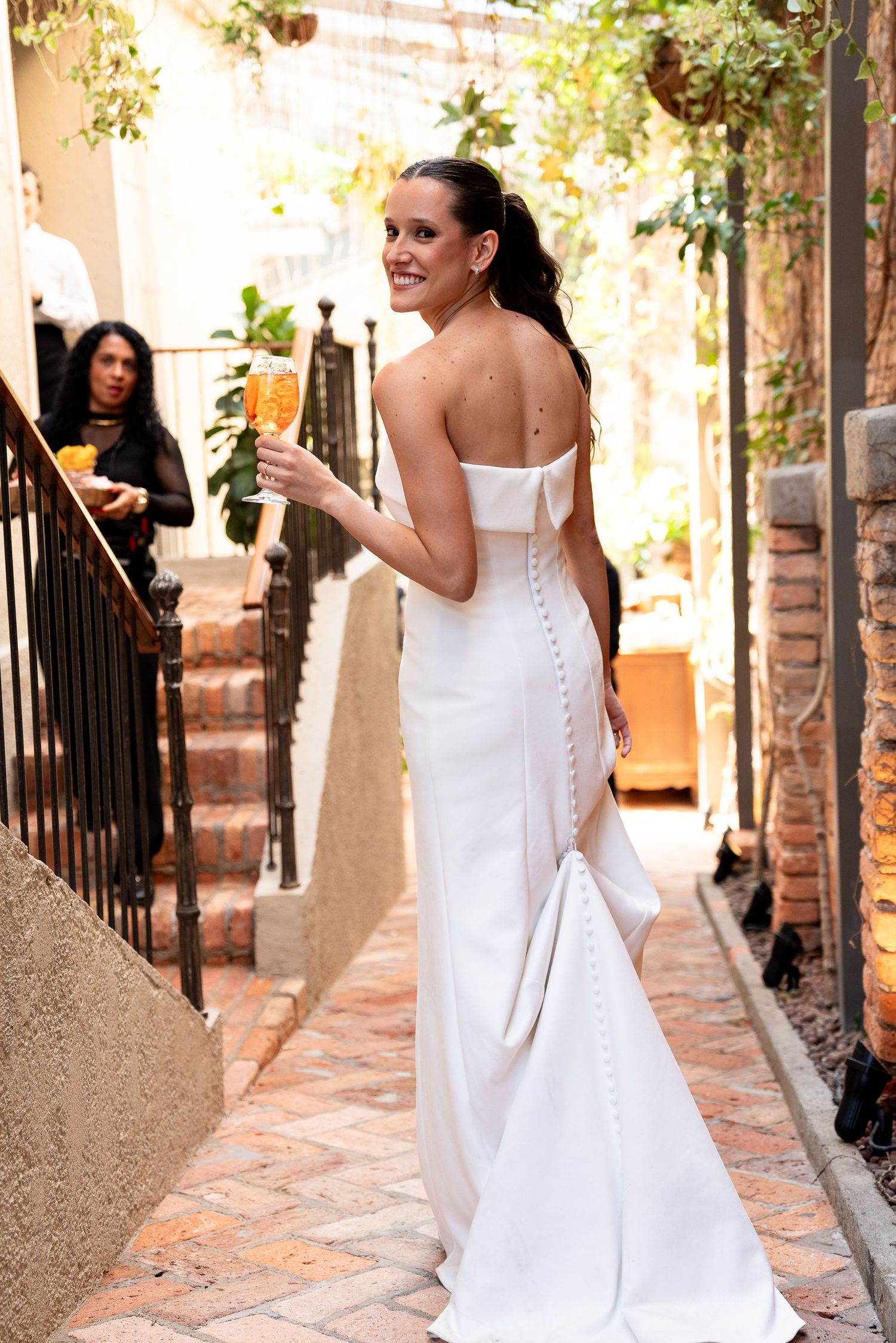 Smiling bride in white strapless gown with bow back detail holds champagne glass at outdoor wedding venue.
