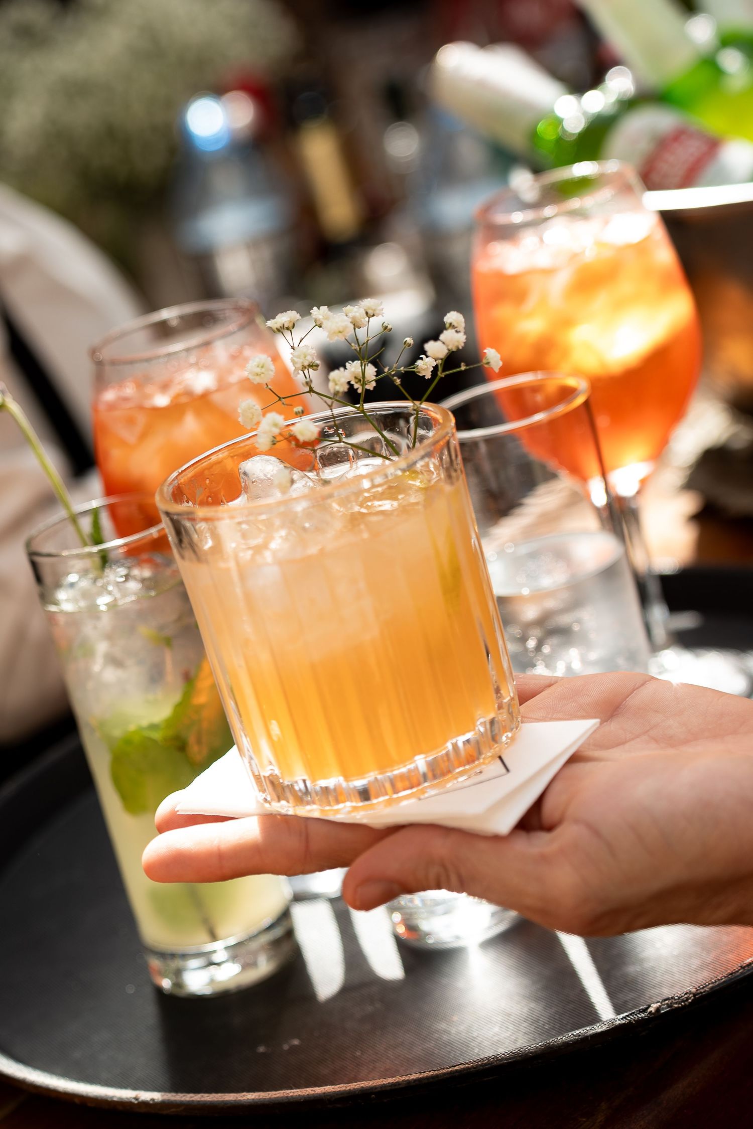A hand holds an amber cocktail garnished with flowers on a tray with assorted colorful drinks in a bar setting.