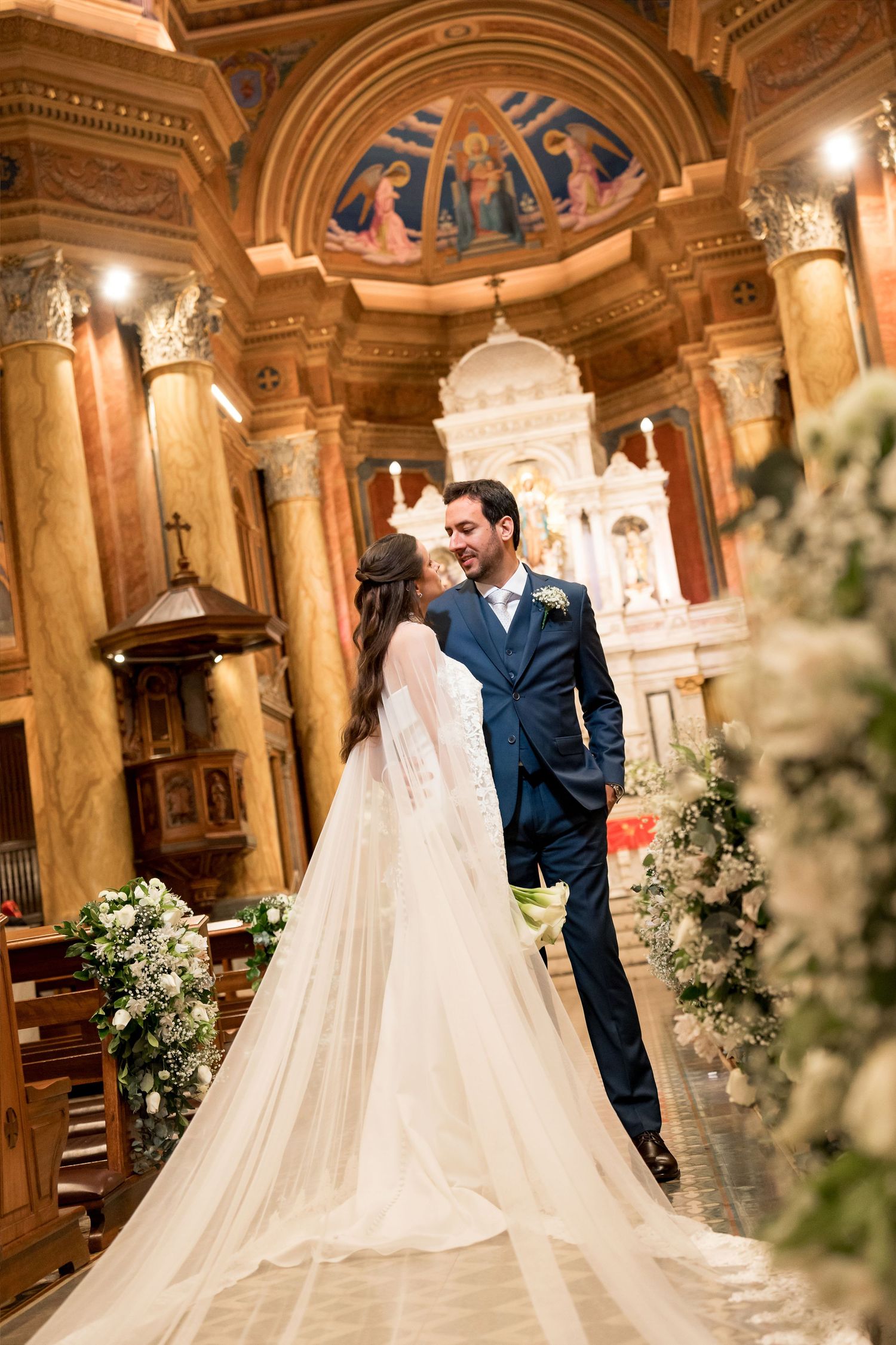 Bride and groom share a romantic moment at the church altar, with ornate columns and religious murals in the background.