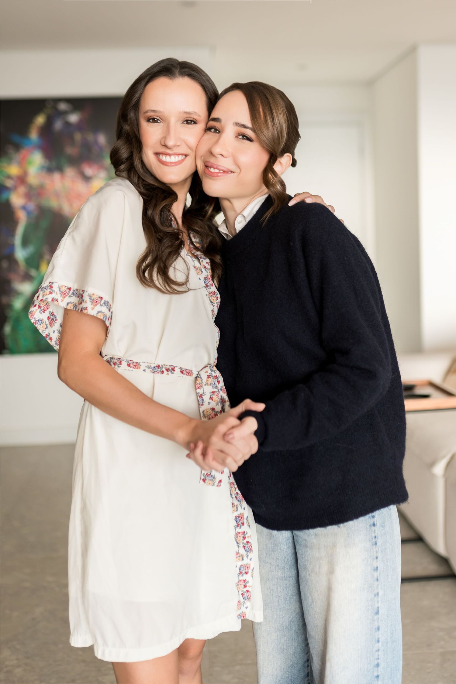 Two smiling women embracing indoors, one in a floral white dress and one in a black sweater with jeans.
