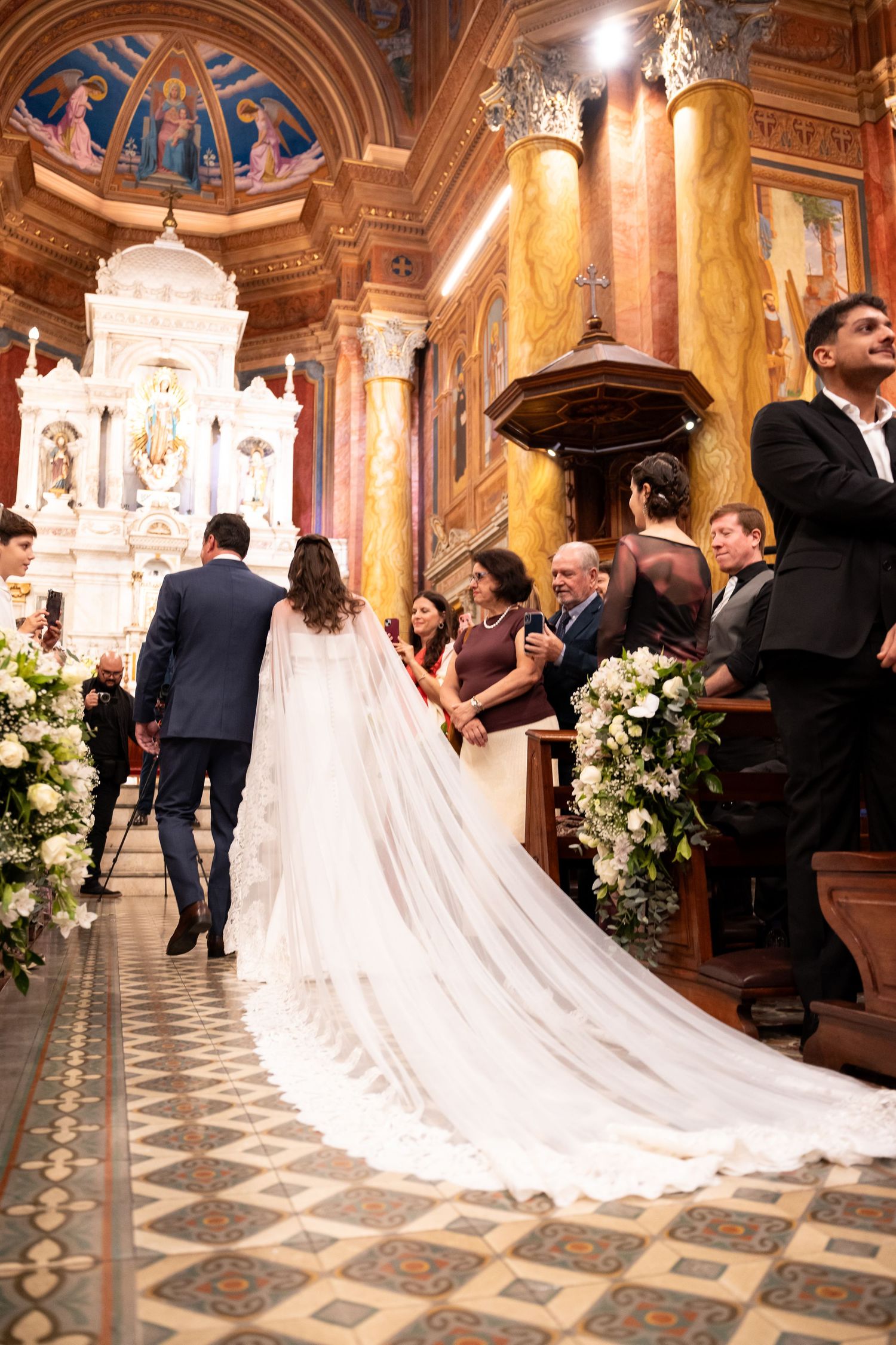 Bride in white gown with long veil walks down ornate church aisle with groom during wedding ceremony.