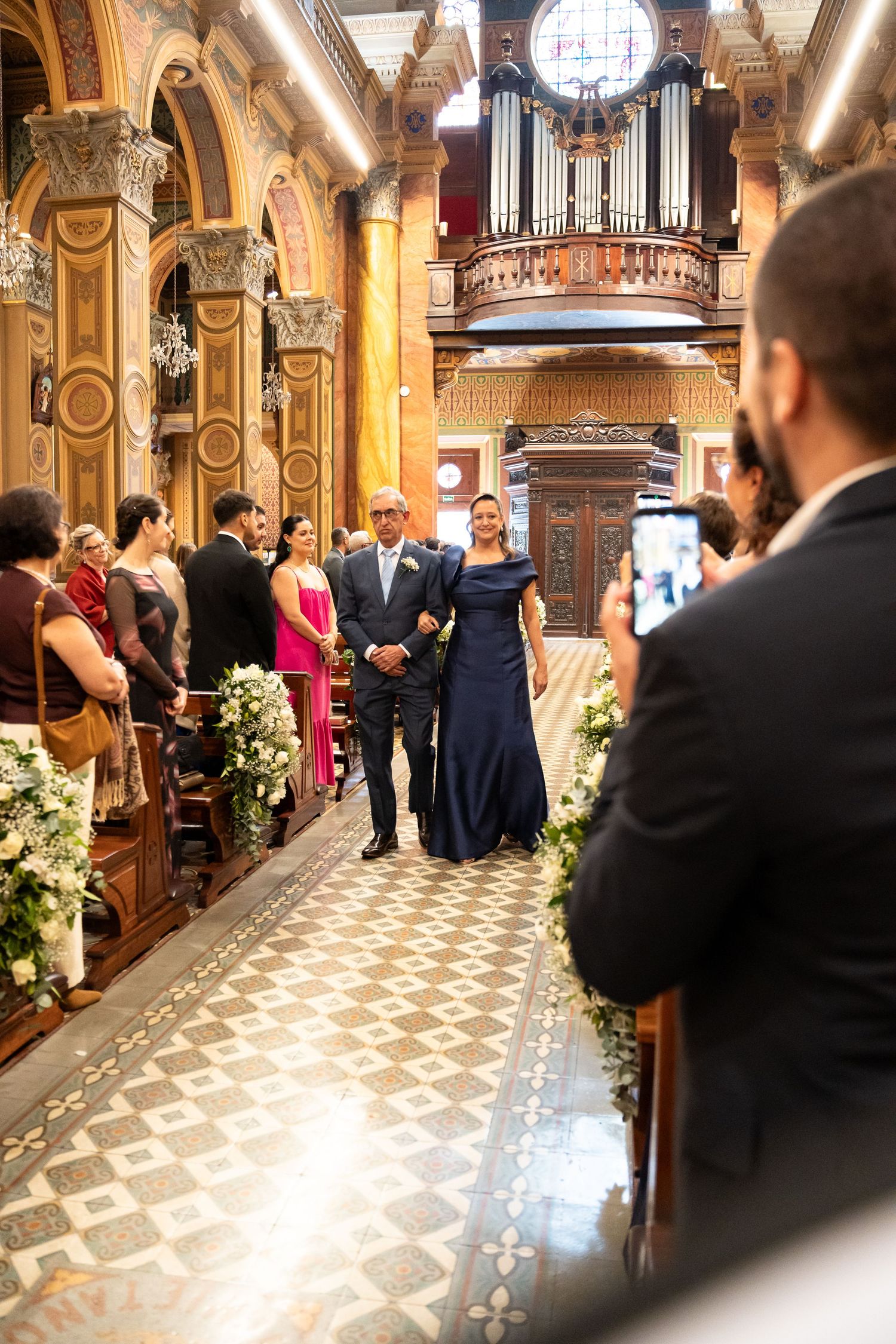 Elegant church wedding ceremony with couple walking down ornate aisle, pipe organ backdrop, floral arrangements, guests watching.