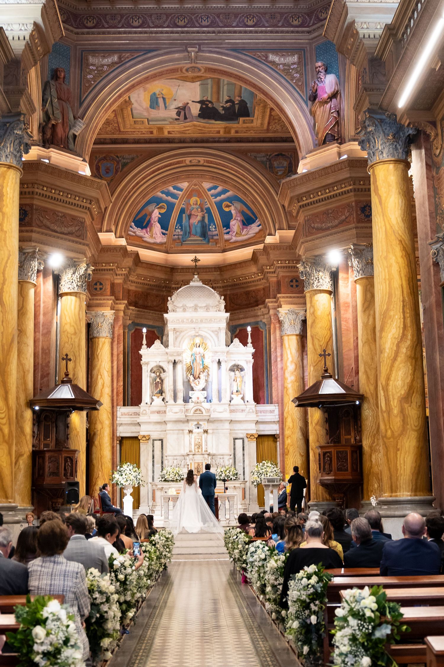 Elegant church wedding ceremony with couple at ornate altar, surrounded by floral arrangements and guests in wooden pews.