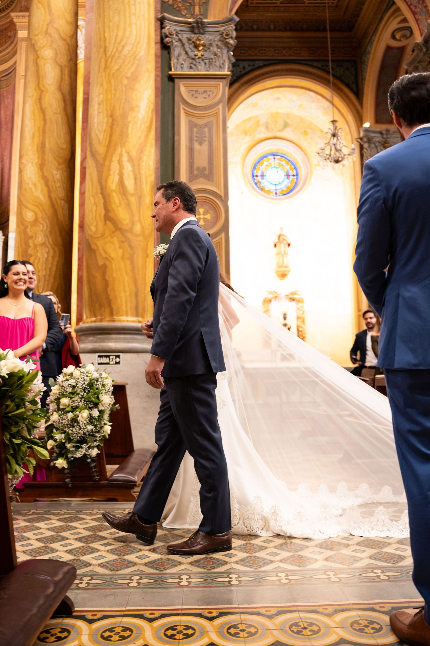Groom walking bride down ornate church aisle, her white veil trailing behind during elegant wedding ceremony.