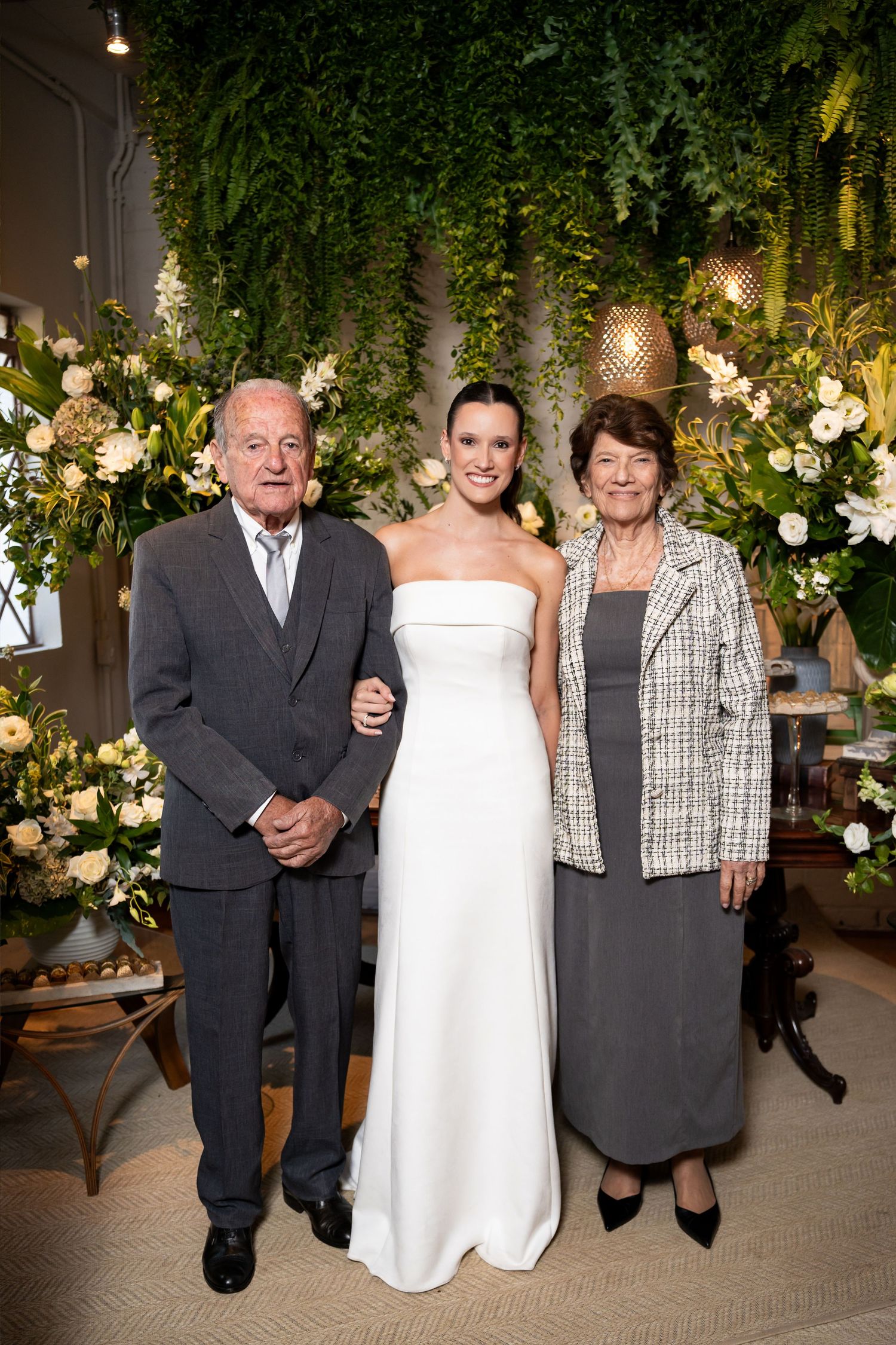 Bride in white gown poses with elderly couple amid lush greenery and white floral arrangements at wedding venue.