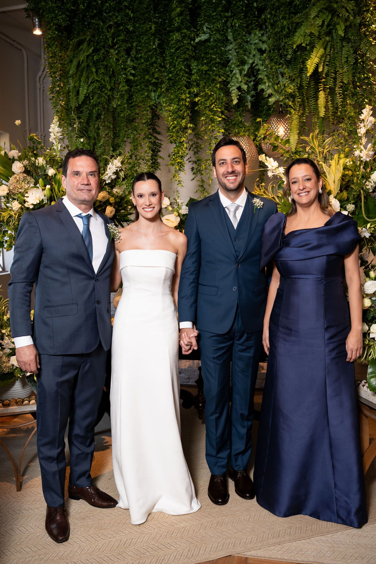 Wedding party of four posing before lush greenery backdrop, bride in white gown, groom in navy suit, flanked by family.