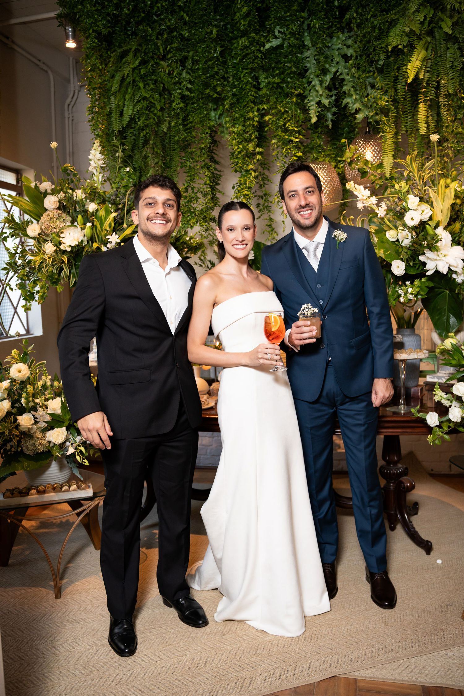 Bride in white gown poses with two men in suits at elegant wedding reception with lush greenery backdrop.