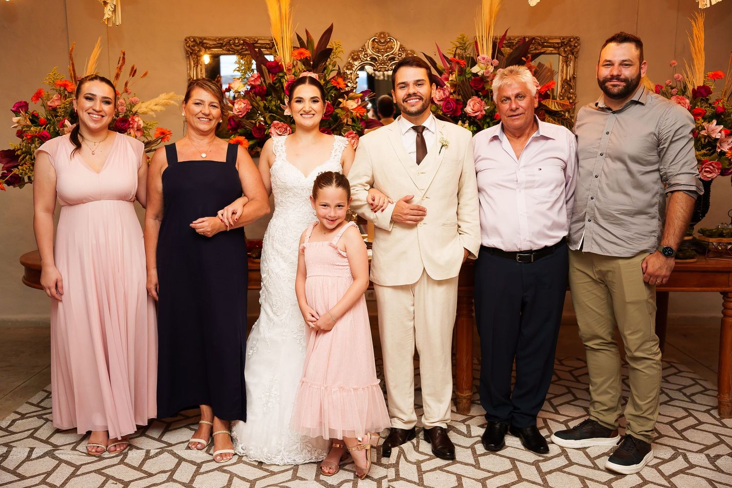 Wedding couple poses with family members in elegant floral venue, bride in white gown, groom in cream suit.