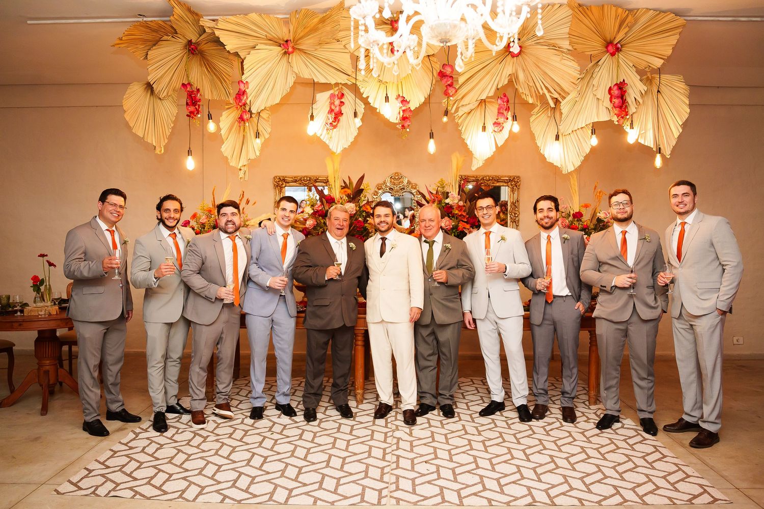 Groom in white suit poses with groomsmen in gray suits and orange ties under floral decorations at wedding venue.