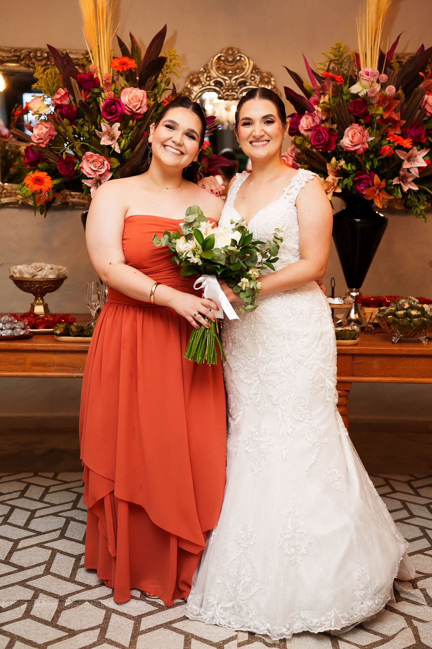 Bride in white lace gown holds bouquet beside bridesmaid in rust orange dress, posing before floral arrangement.