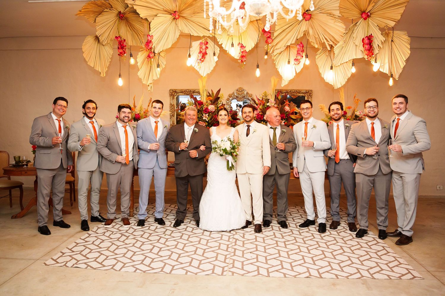 Wedding party poses indoors with bride and groom, groomsmen in gray suits with orange ties, beneath floral ceiling decor.